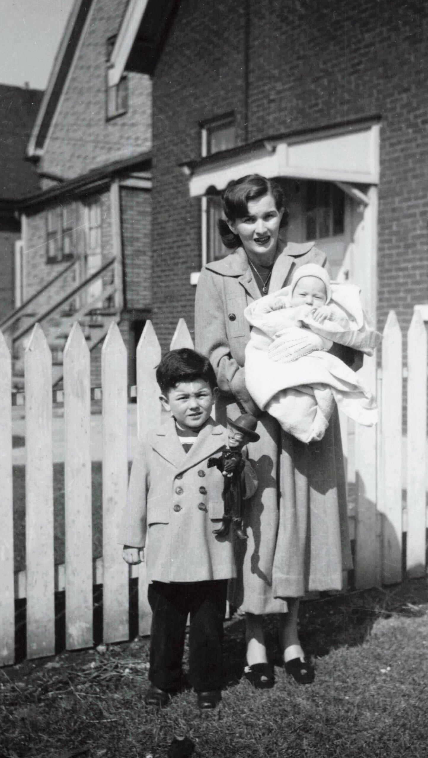 Mother holding a baby wrapped in a blanket, standing beside a white picket fence with a young boy beside her
