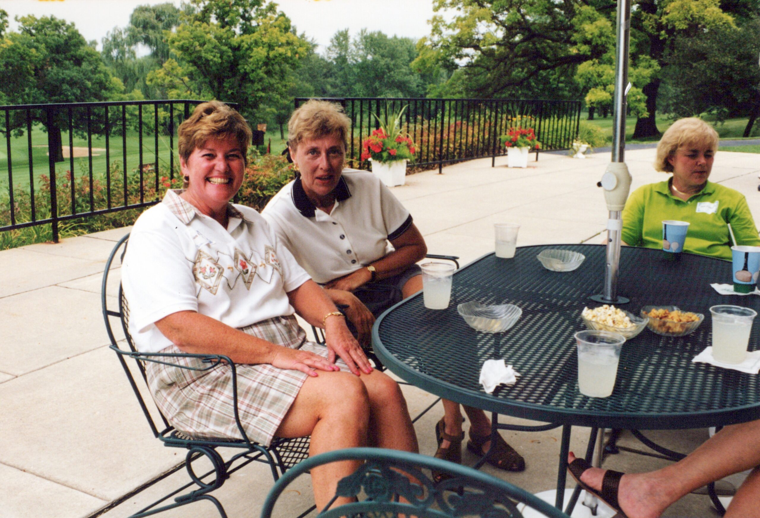 Three women sit around a metal patio table outdoors, enjoying snacks and drinks in a park setting.