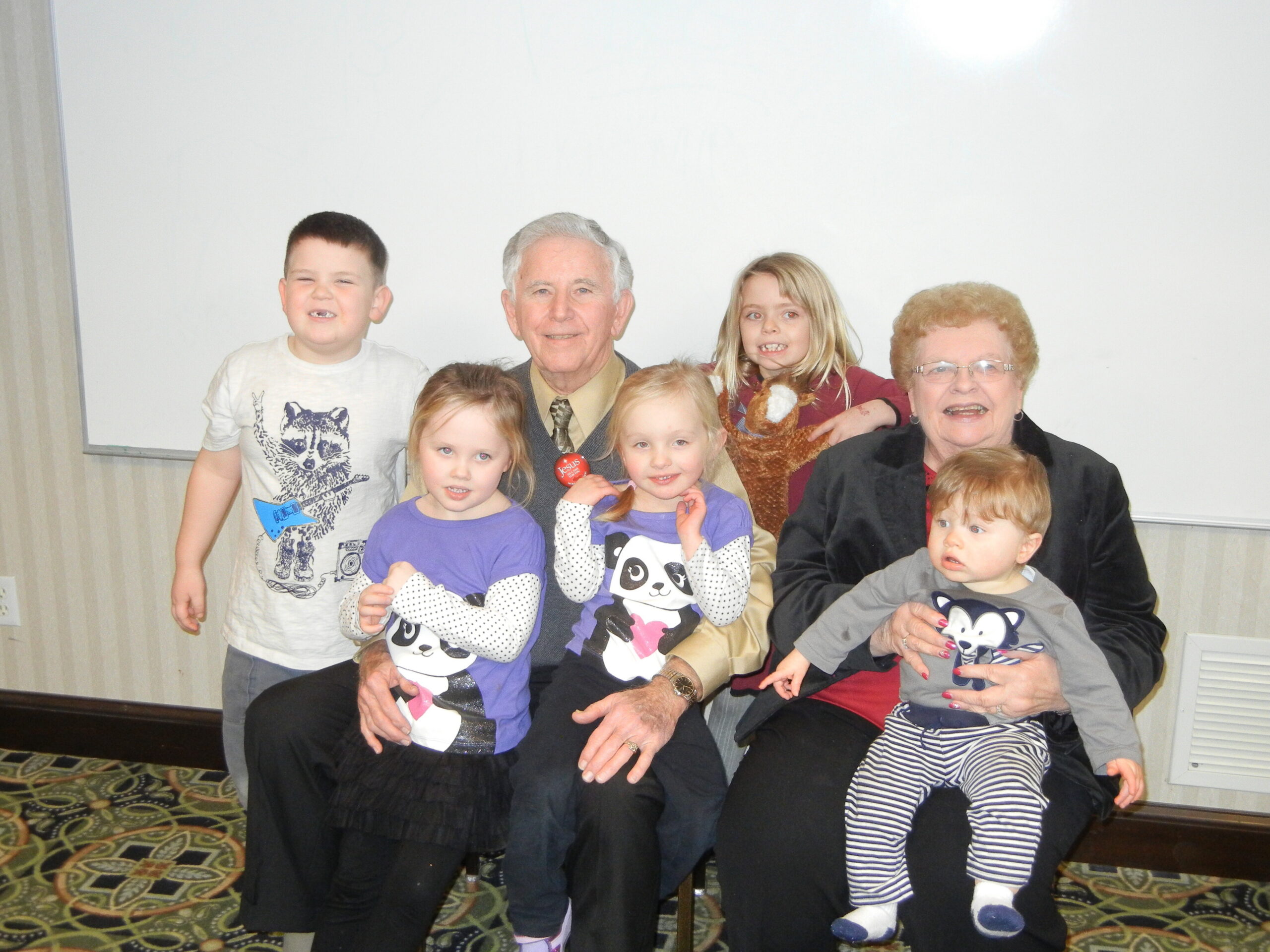 Group photo of an elderly couple surrounded by smiling children indoors, posing for a family portrait.