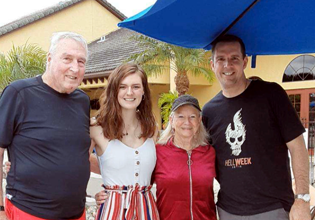 Four adults smiling together for a casual outdoor group photo under a blue umbrella at a sunny venue; palm trees in the background.