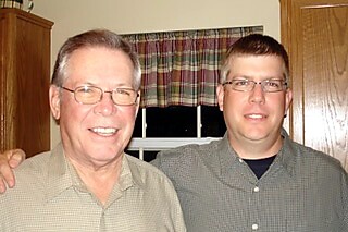 Two men smiling at the camera indoors, standing close together with wooden cabinets behind them.
