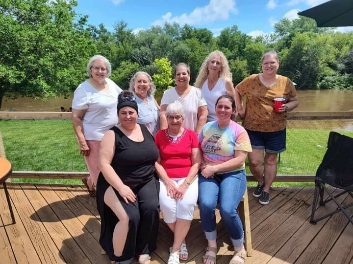Nine women posing together on a wooden deck by a river, smiling with trees and blue sky in the background.