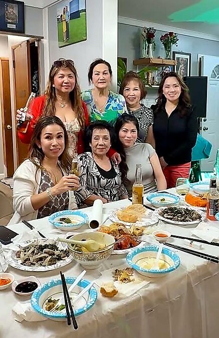 Group of seven women smiling around a dining table with assorted dishes and drinks in a home setting, posing for a photo.