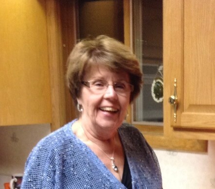 Smiling woman with short brown hair and glasses wearing a blue knit sweater in a kitchen with wooden cabinets nearby.
