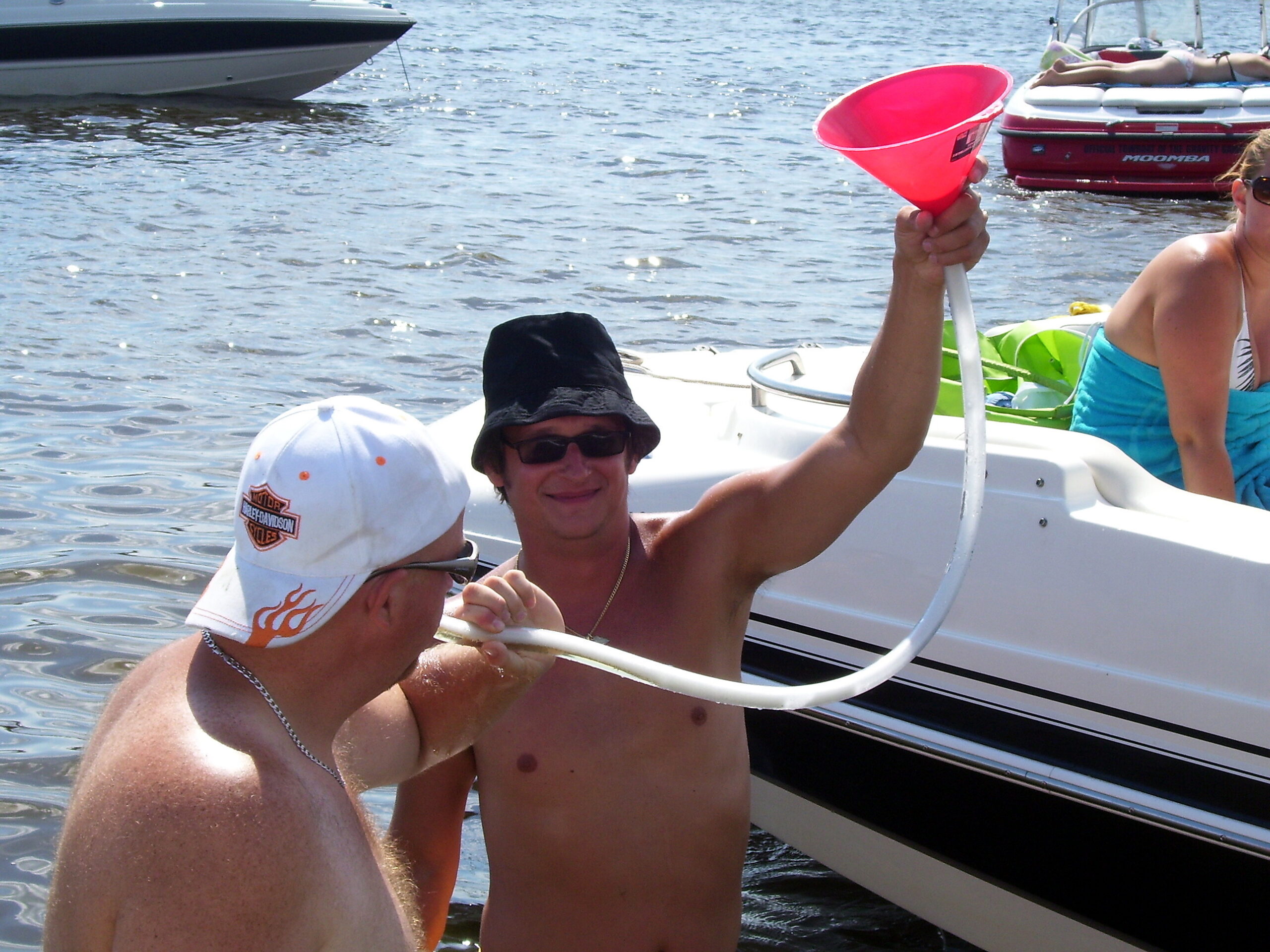 Three people on a white boat on sparkling water; a man in a black hat and sunglasses, another shirtless man, and a woman in a blue swimsuit holding a bright pink funnel/megaphone-like object.