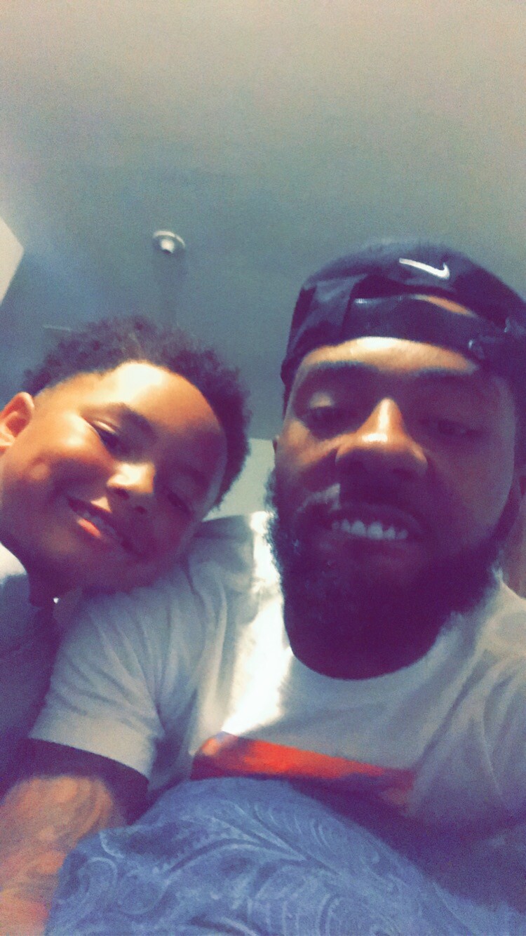 A man and a child smiling for a close-up selfie in bed, with the child resting his head on the man's shoulder as they lean in together.