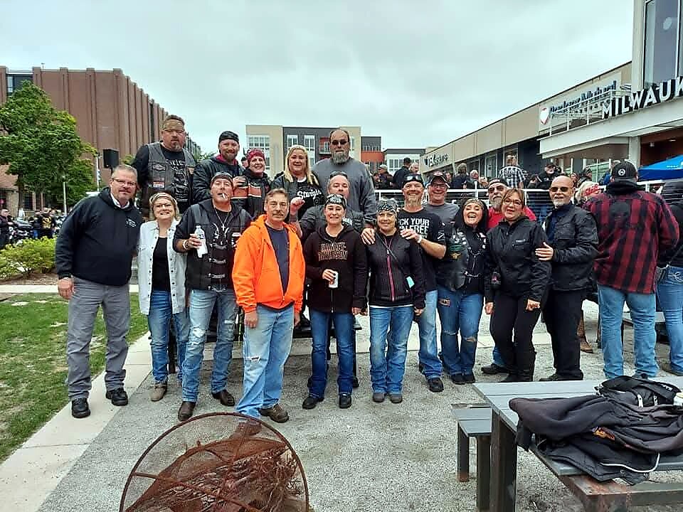 Group of motorcycle club members posing together outdoors in a plaza, many wearing leather jackets and jeans, smiling at the camera.
