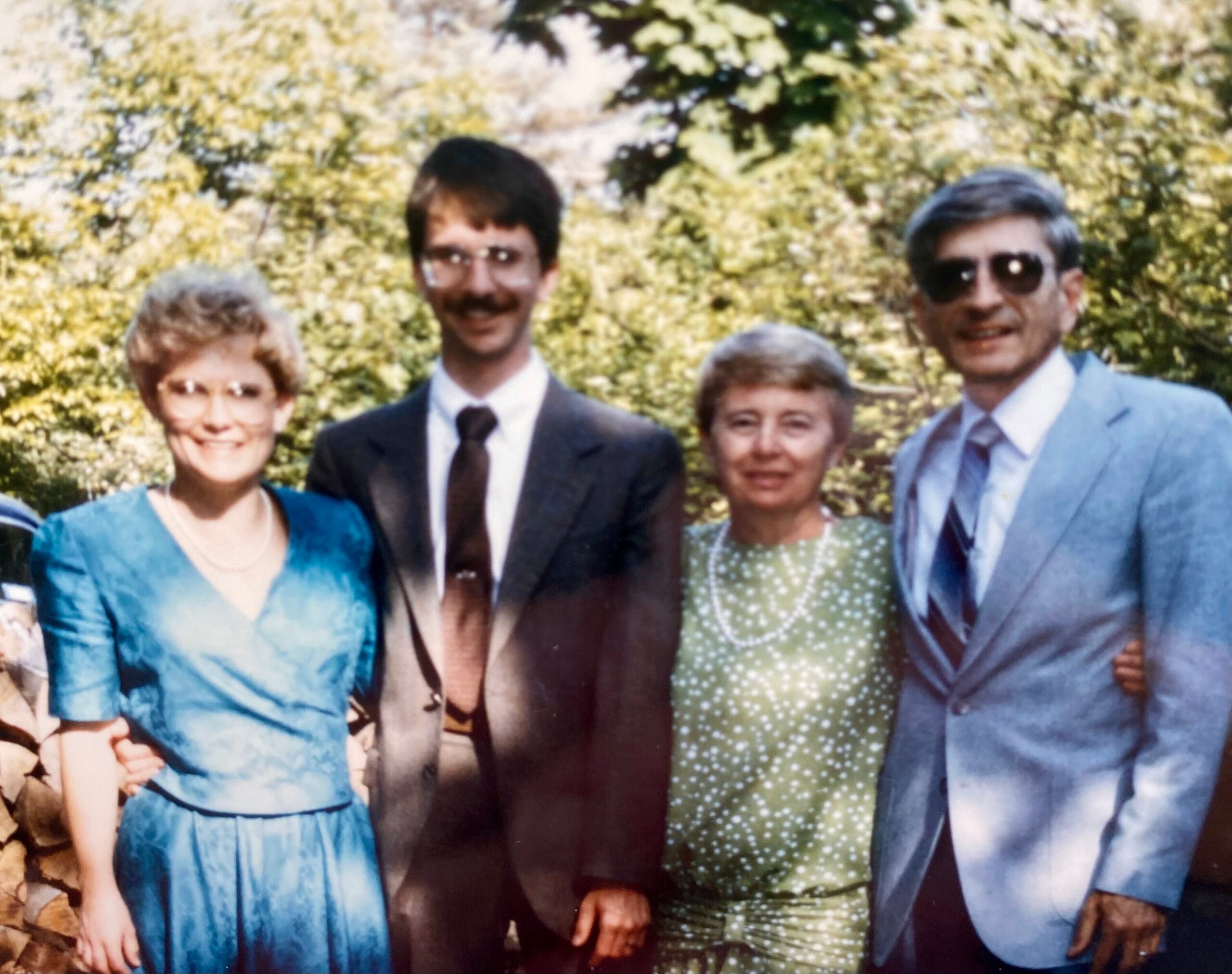 A small group of four adults posing outdoors in vintage clothing, smiling for the photo in a sunny garden setting.