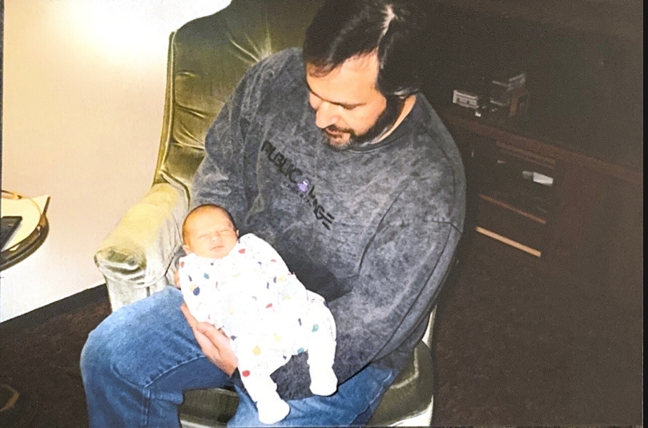 Man with a beard seated in a green armchair, holding a newborn baby wrapped in a colorful blanket.