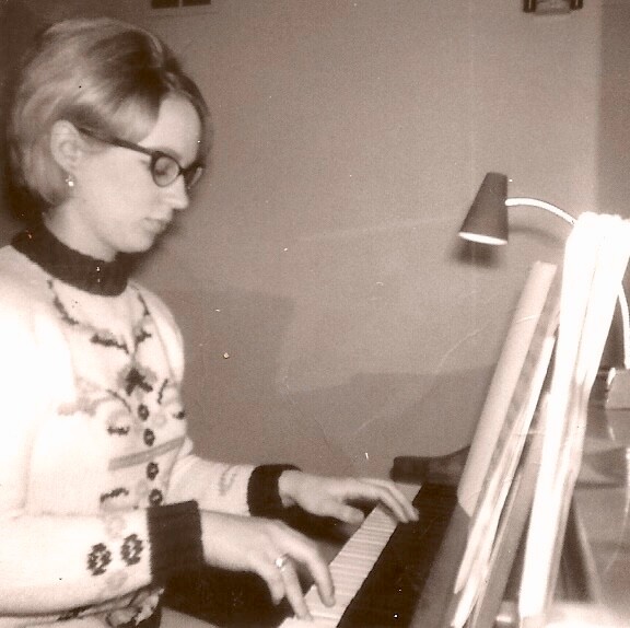 Woman wearing glasses and a patterned sweater plays a piano under a desk lamp.