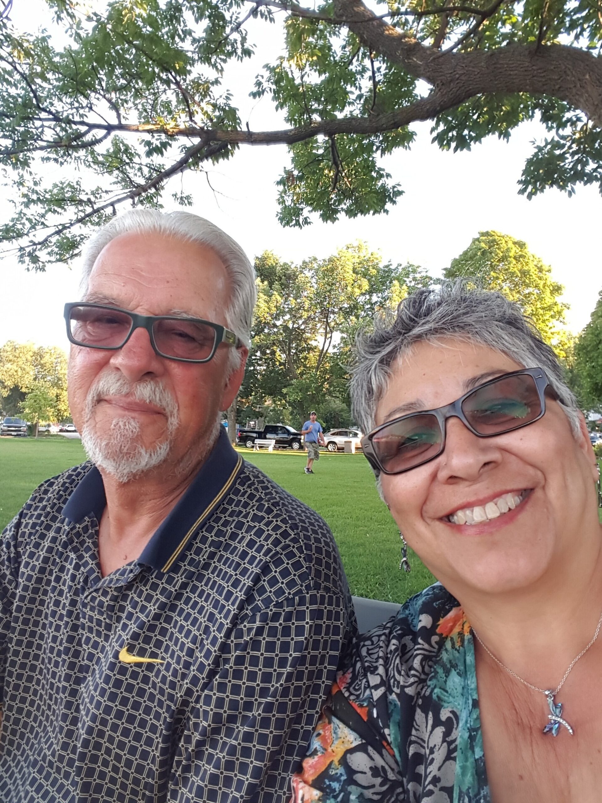 Two adults take a sunny park selfie; man with glasses on the left and a smiling woman with short grey hair and sunglasses on the right, green trees in the background.