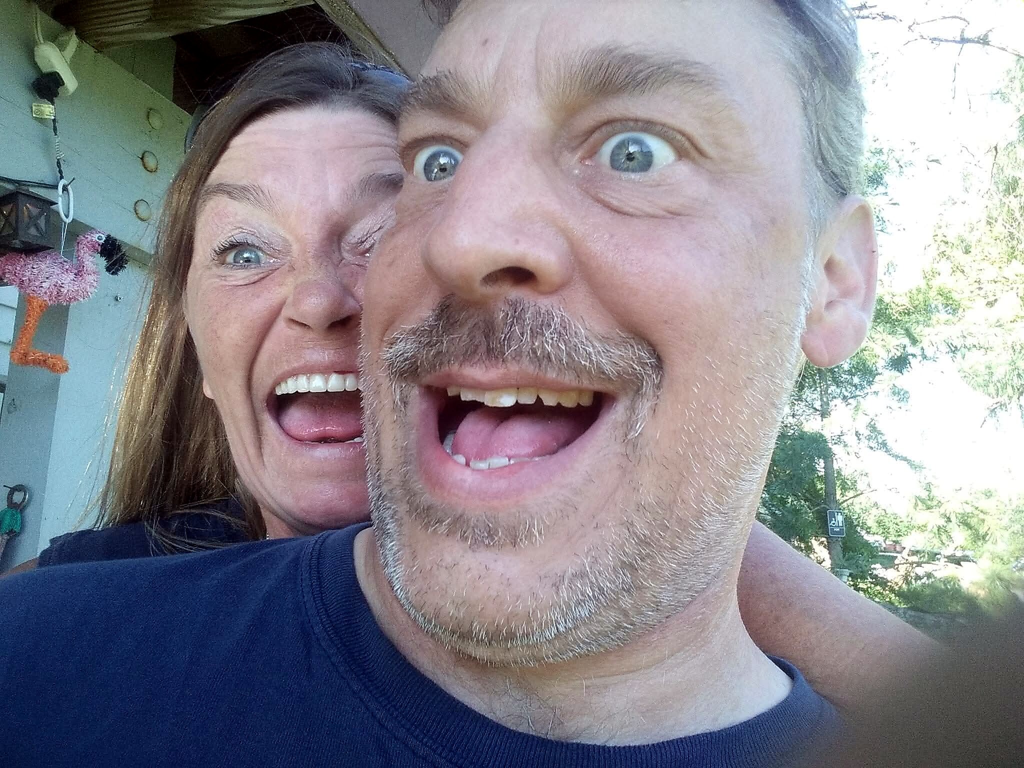 Close-up of a smiling couple taking a goofy selfie outdoors, faces pressed together and mouths open in laughter.
