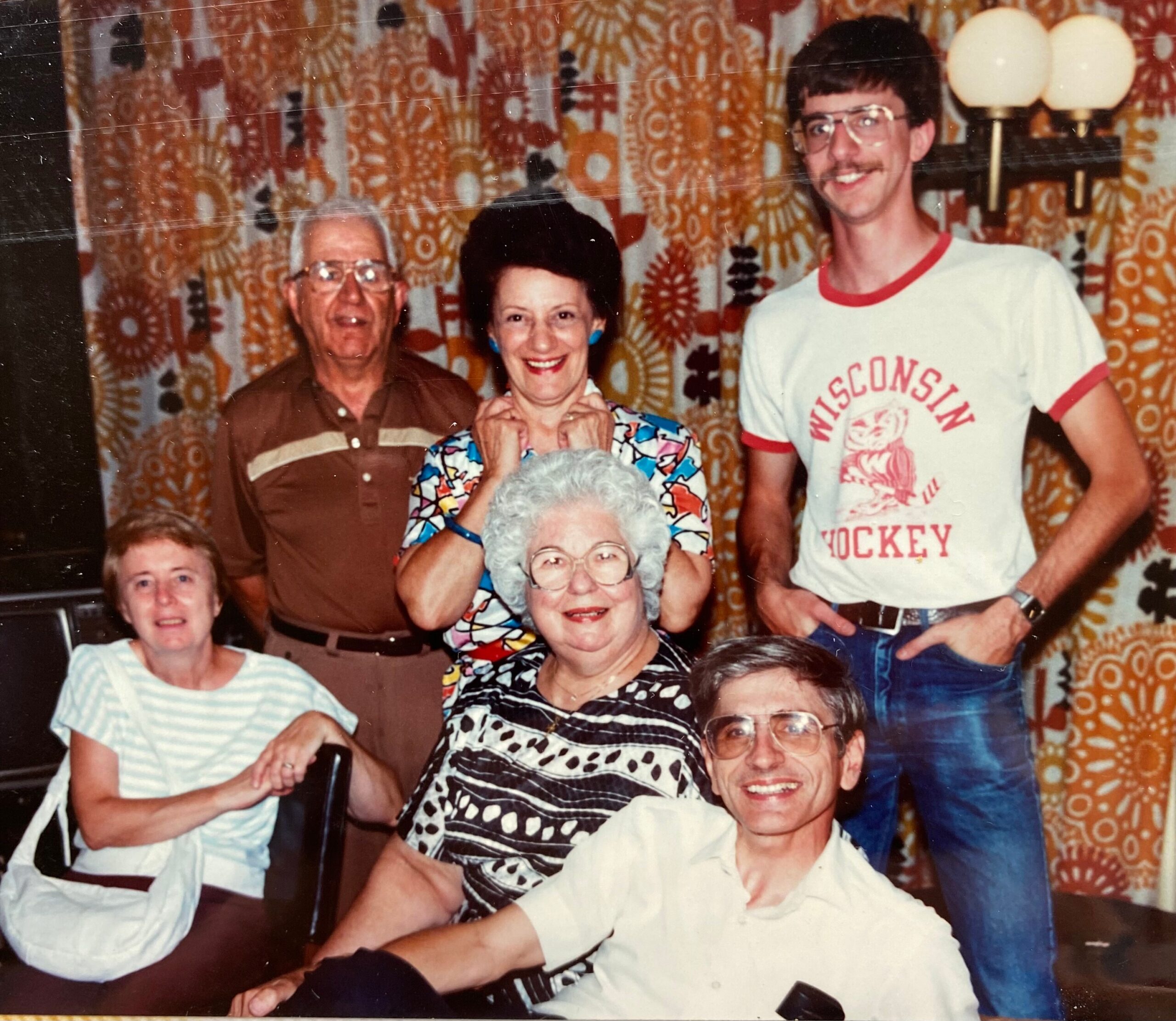 Group of smiling adults posing for a photo in a room with bold orange and brown patterned curtains; a woman in a colorful blouse stands center, flanked by others.