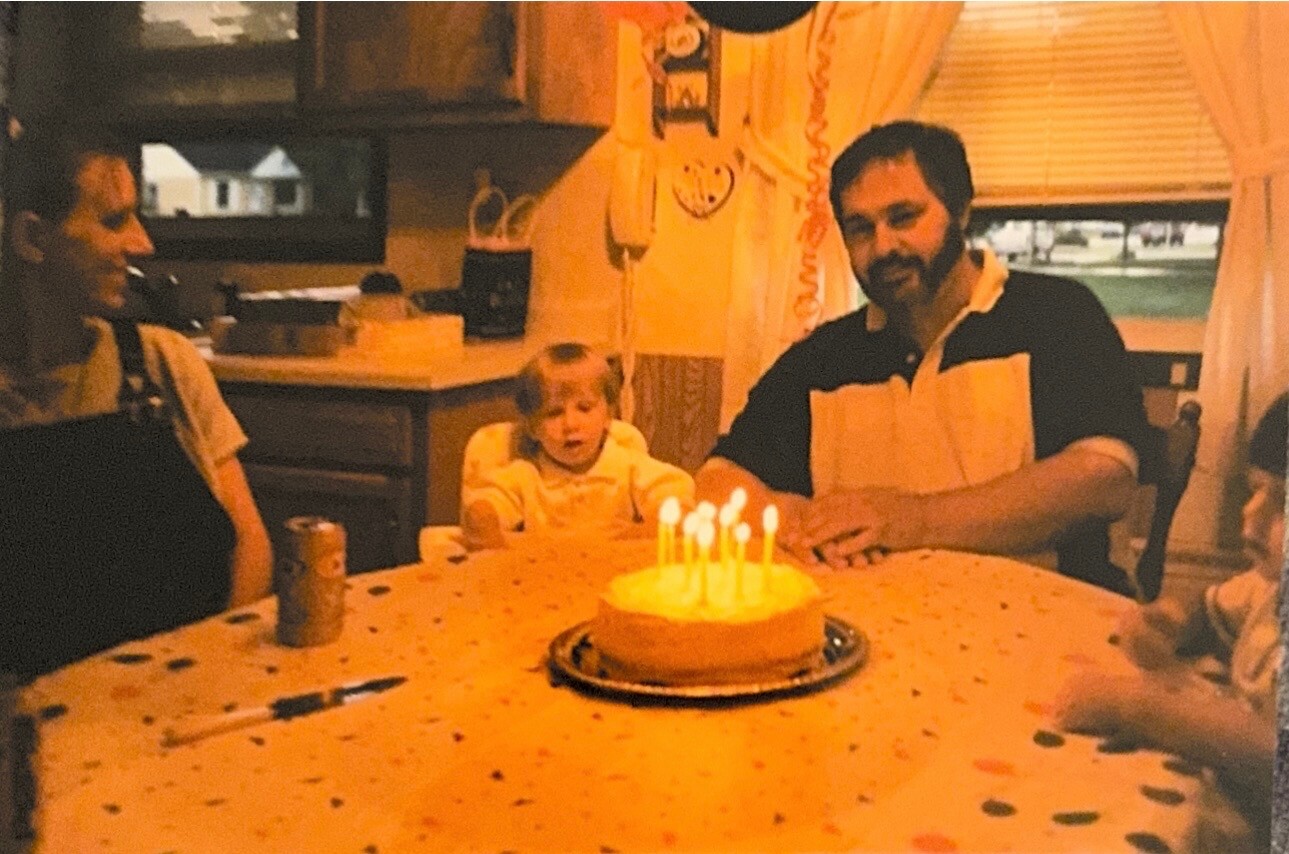 A child and two adults sit around a table with a lit birthday cake indoors, celebrating a birthday.