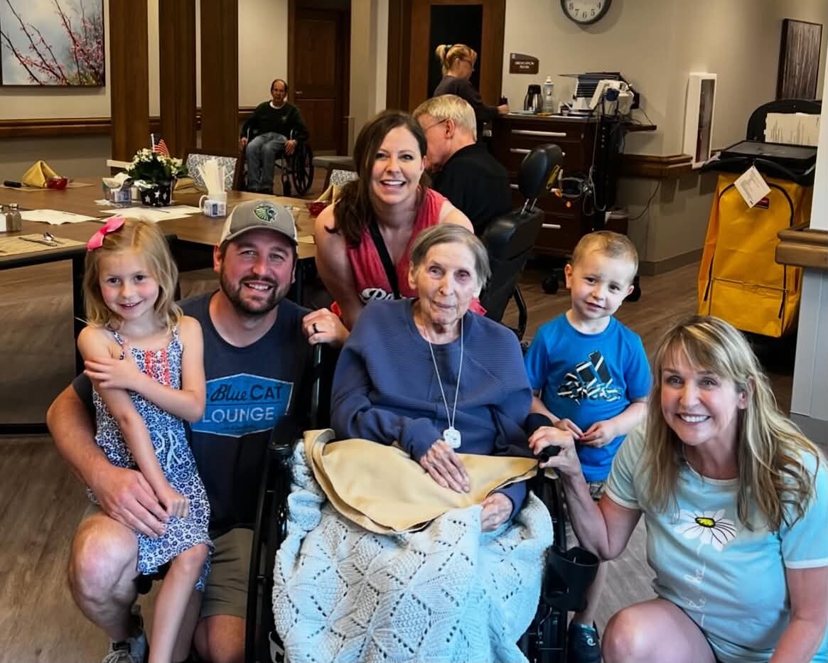 Family posing with an elderly woman in a wheelchair in a bright reception area of a care facility.
