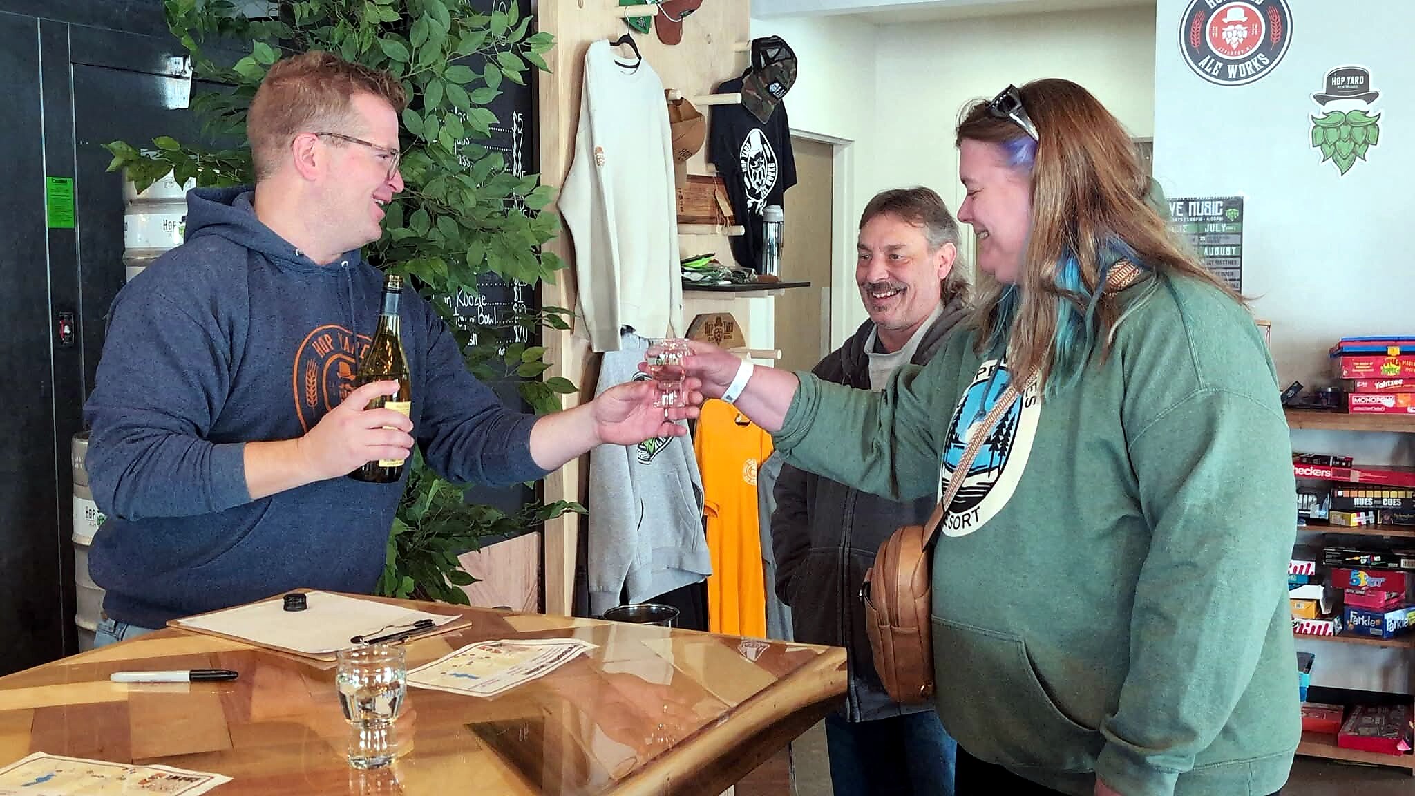 Man in a blue hoodie hands a beer to a woman in a green jacket across a wooden counter as others smile in the background toasting.