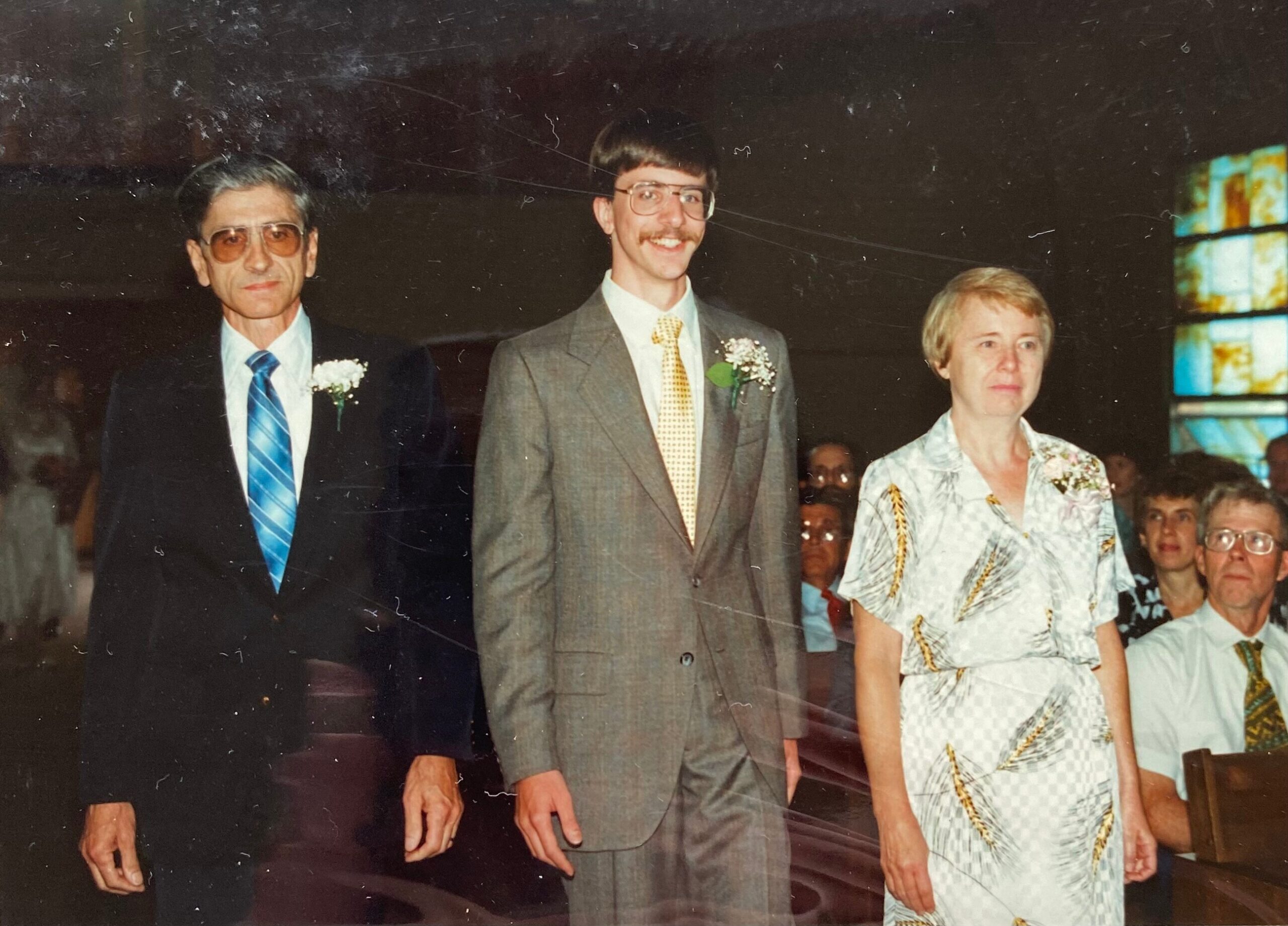 Three adults in formal attire standing together at a ceremony: an older man in a dark suit with blue tie, a younger man in a gray suit with yellow tie and boutonniere, and a woman in a light patterned dress on the right.