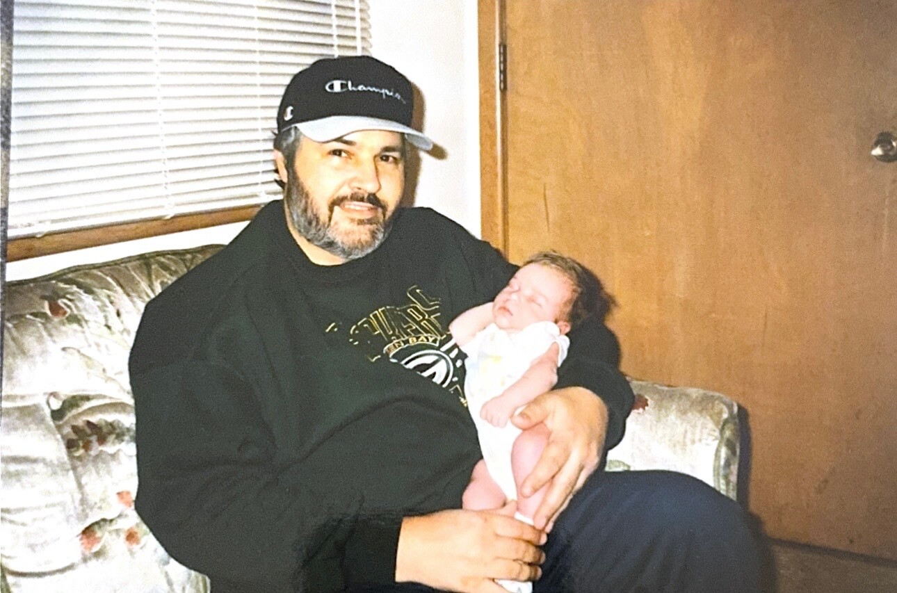 Man with a beard and baseball cap sitting on a floral couch, cradling a sleeping newborn baby in white sleepsuit.