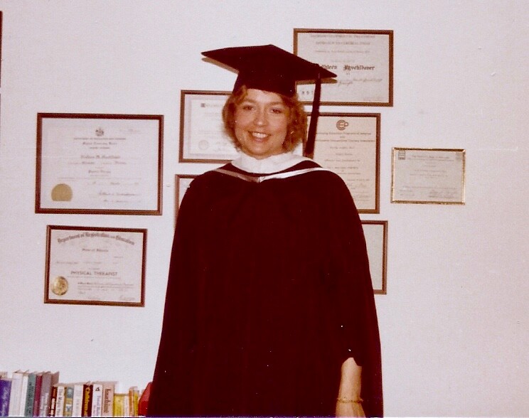 Graduate in cap and gown posing in front of framed certificates on the wall.