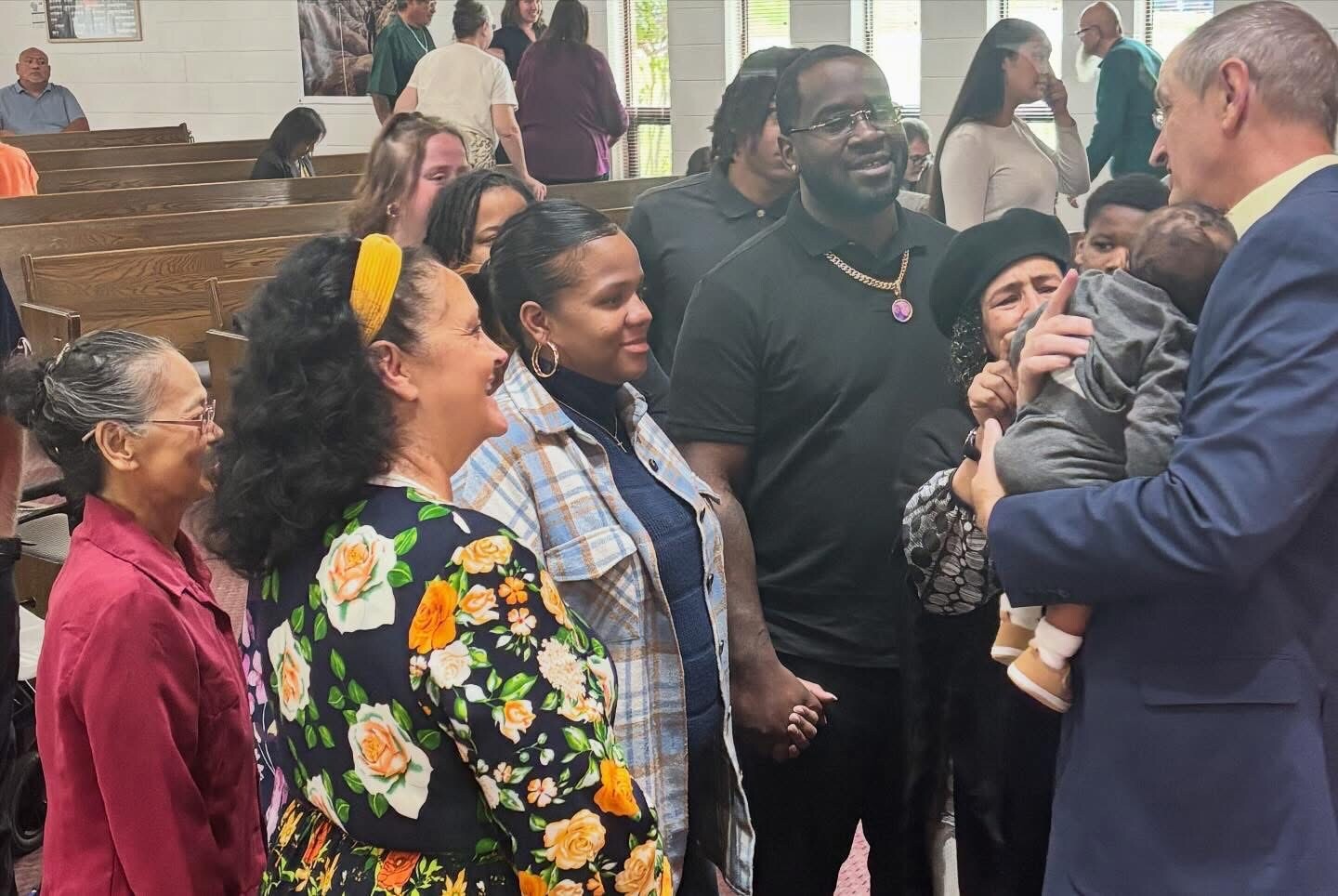 Diverse group of adults in a church hall, smiling as a man in a blue suit holds and presents a baby to a woman.