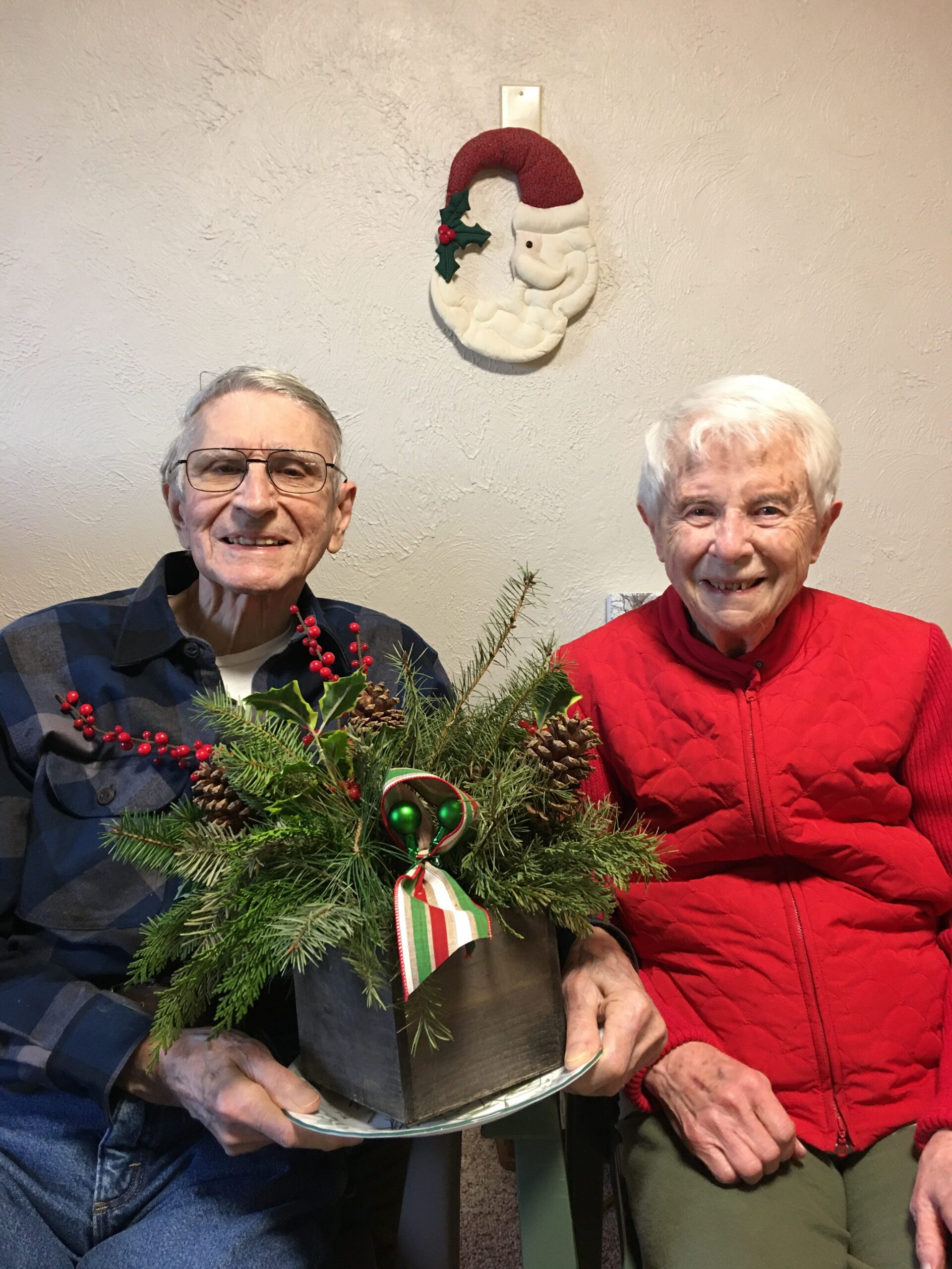 Two elderly people sit smiling, holding a festive evergreen centerpiece with pinecones and red berries in a black container in front of a wall with a Santa wreath above them.