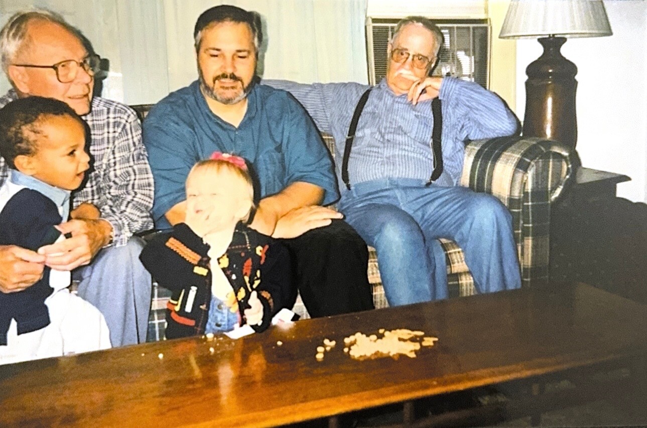 Family on a plaid sofa: an older man holds a toddler, a man in blue sits center, a man with suspenders sits to the right; a girl with a pink bow sits in front, crackers on the table.