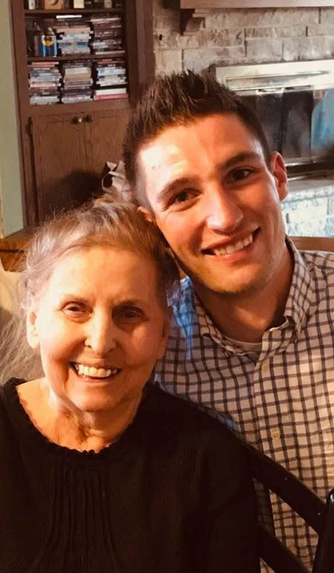 Smiling elderly woman and younger man posing together indoors, warm home setting with a stone fireplace in the background.
