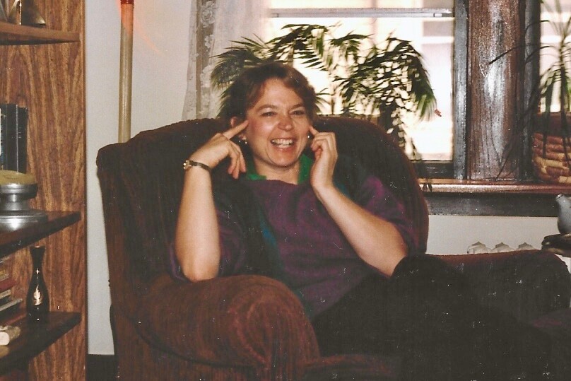Woman relaxing in a brown armchair, smiling while talking on a home landline cordless phone in a cozy living room with a plant behind her.