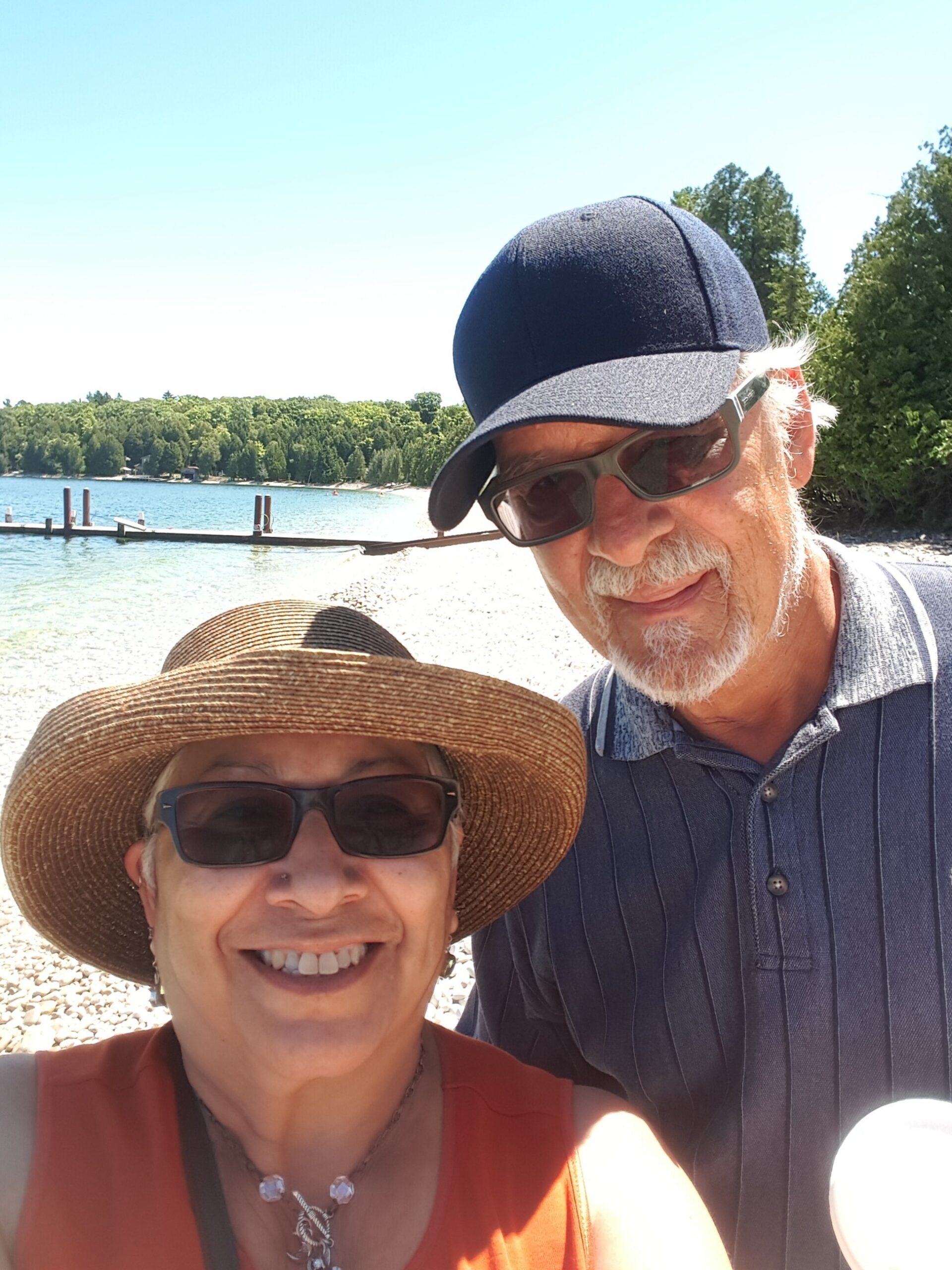 Couple taking a sunny lakeside selfie; woman wears a wide-brim hat and sunglasses, man beside her wearing a dark cap and striped shirt by the water.