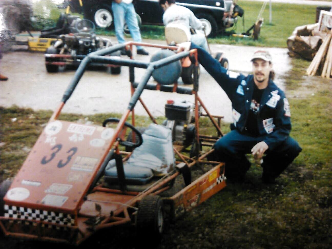 Man in a patched jacket kneels beside a red go-kart with the number 33, in a yard with tools and people in the background.