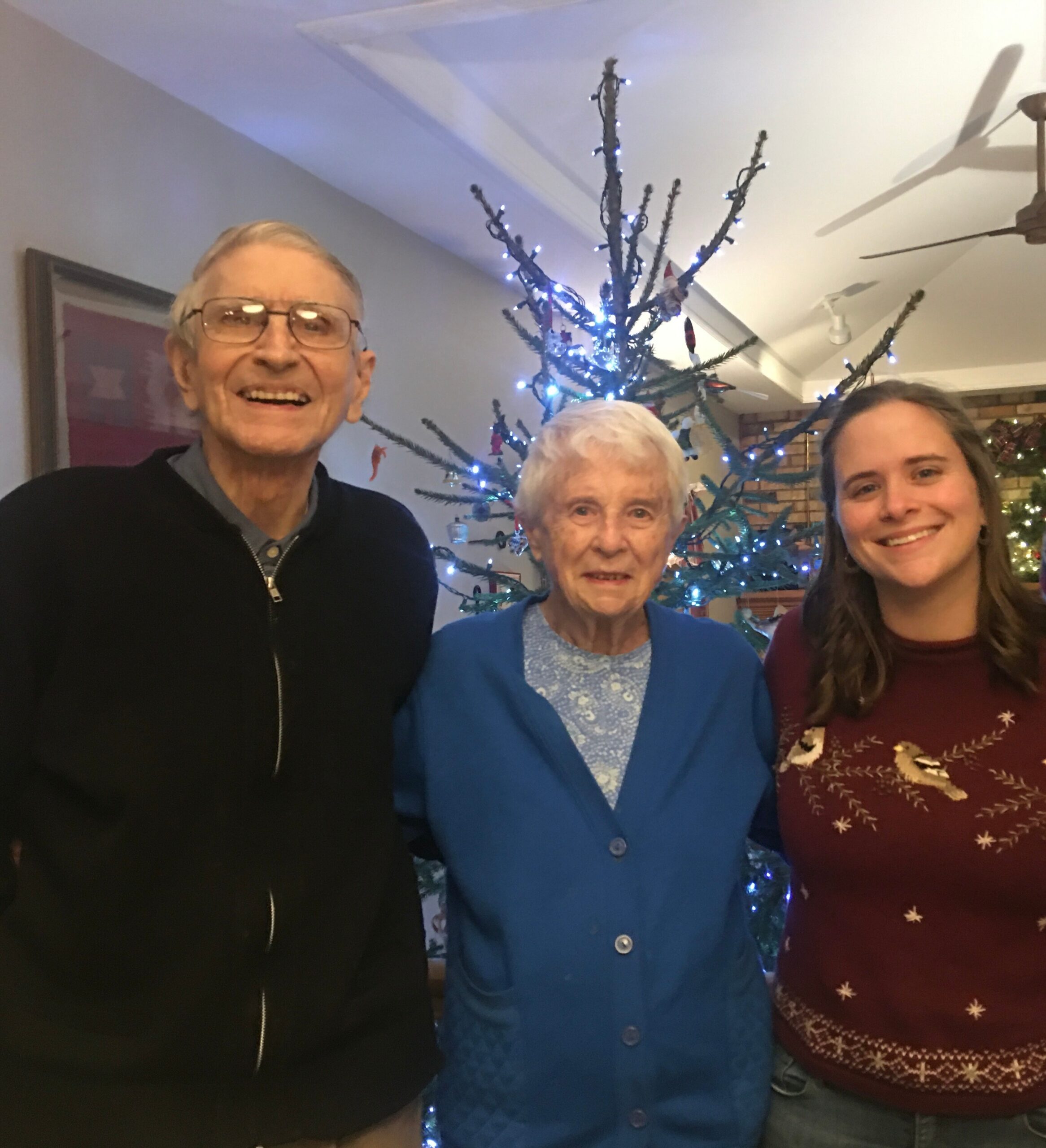 Three people pose for a selfie indoors in front of a lit Christmas tree and colorful lights, smiling at the camera.
