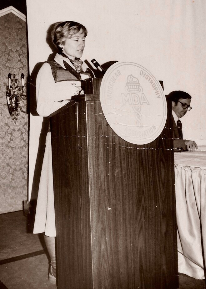 Woman speaking at a podium with a large MDA logo, microphone in hand, while a man sits at a table in the background (black-and-white photo).