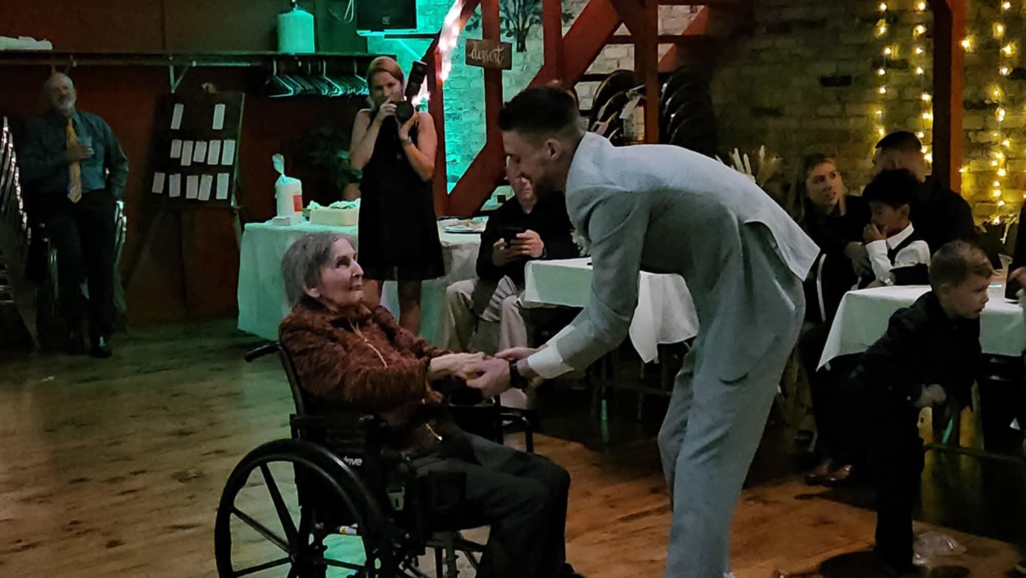 An elderly woman in a wheelchair being greeted and shaken hands with a man in a light suit at a festive indoor gathering.