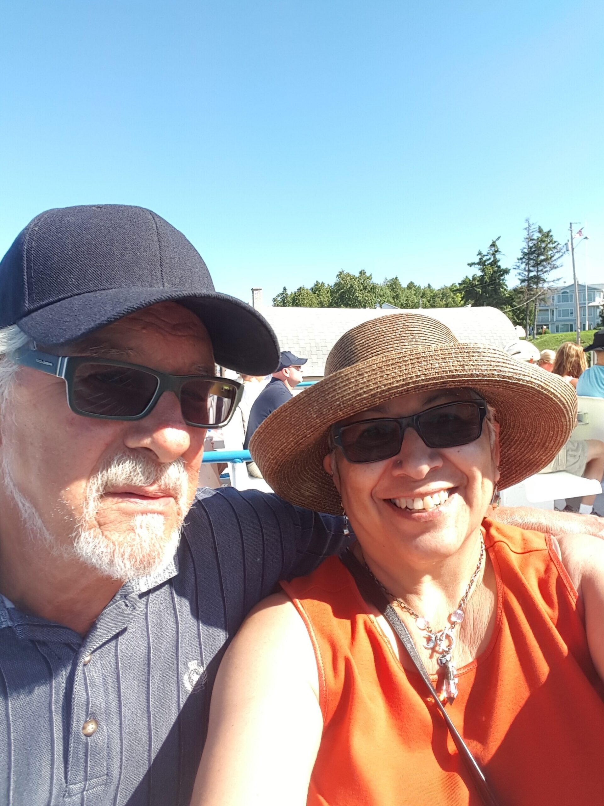 Two smiling people pose for a sunny outdoor selfie; man in a dark cap and sunglasses, woman in a straw hat and sunglasses nearby.