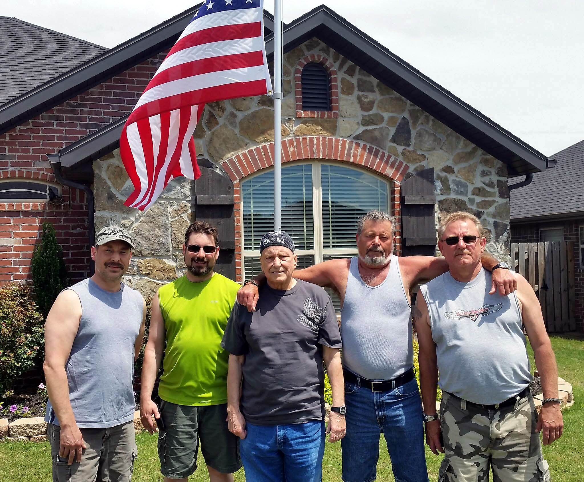 Group of men posing in front of a stone house with an American flag on a pole