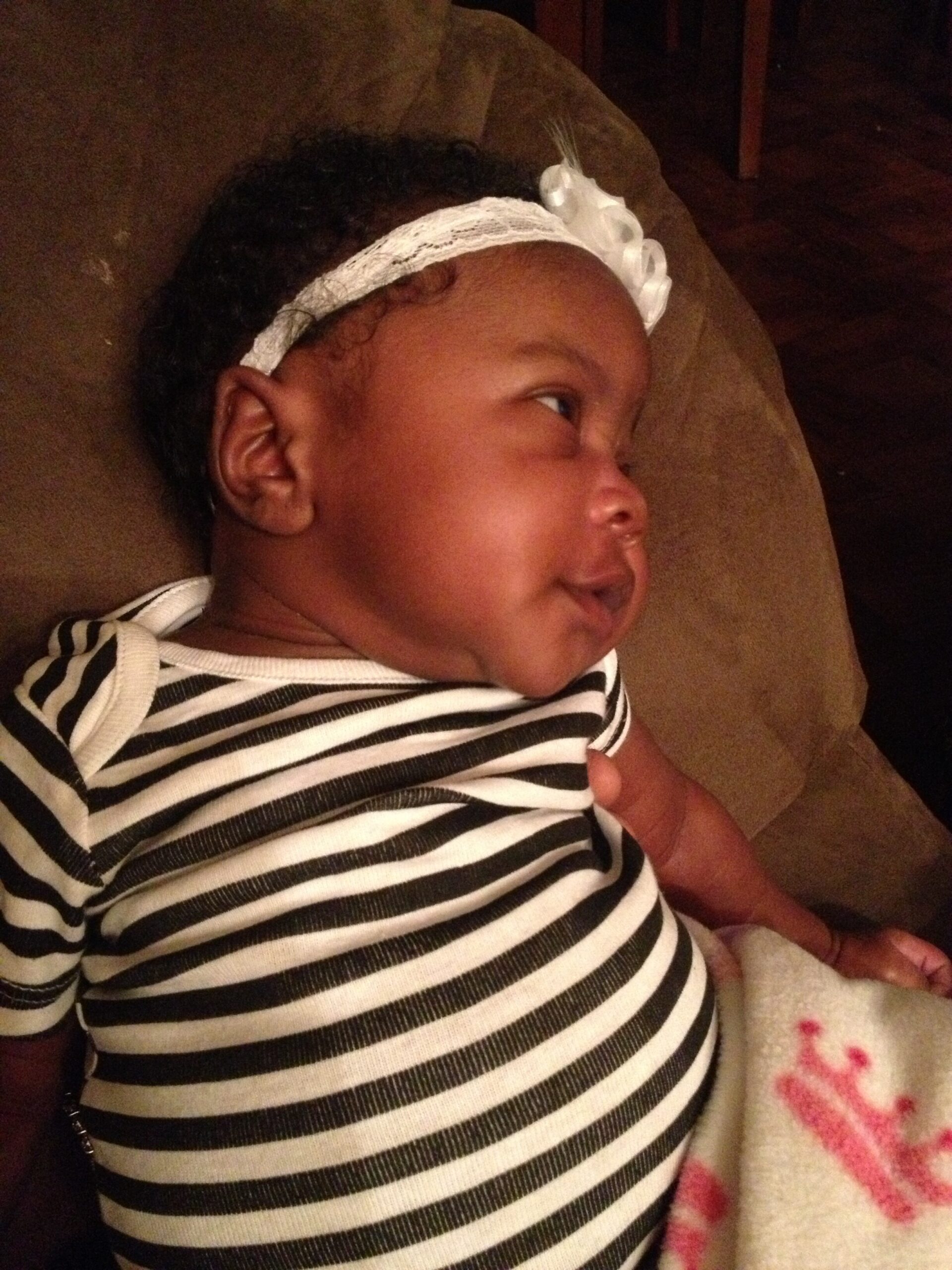 Infant lying on a brown couch, wearing a black-and-white striped outfit and a white lace headband.