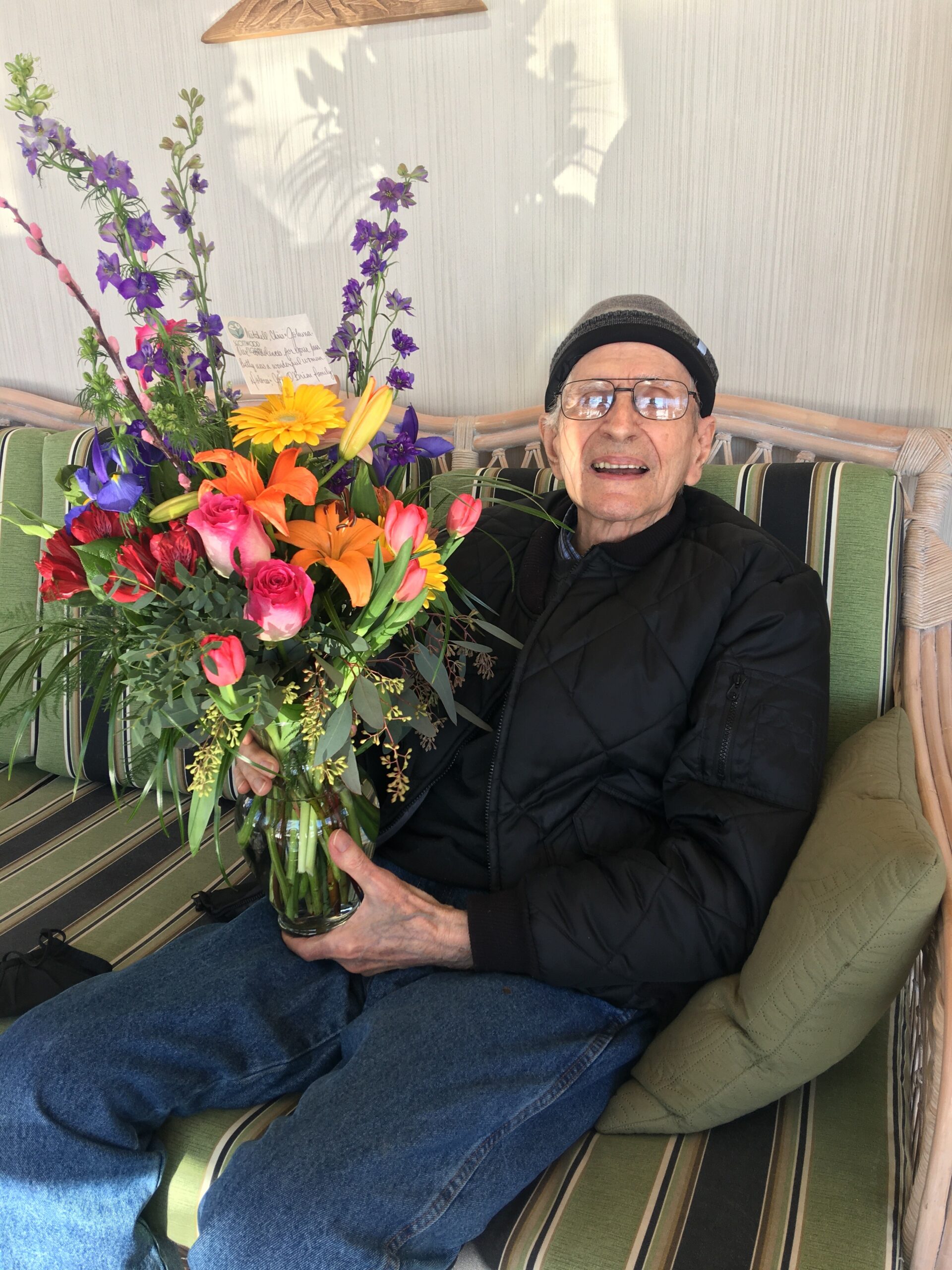 Elderly man sitting on a striped green outdoor couch holding a large colorful bouquet of flowers and smiling at the camera.