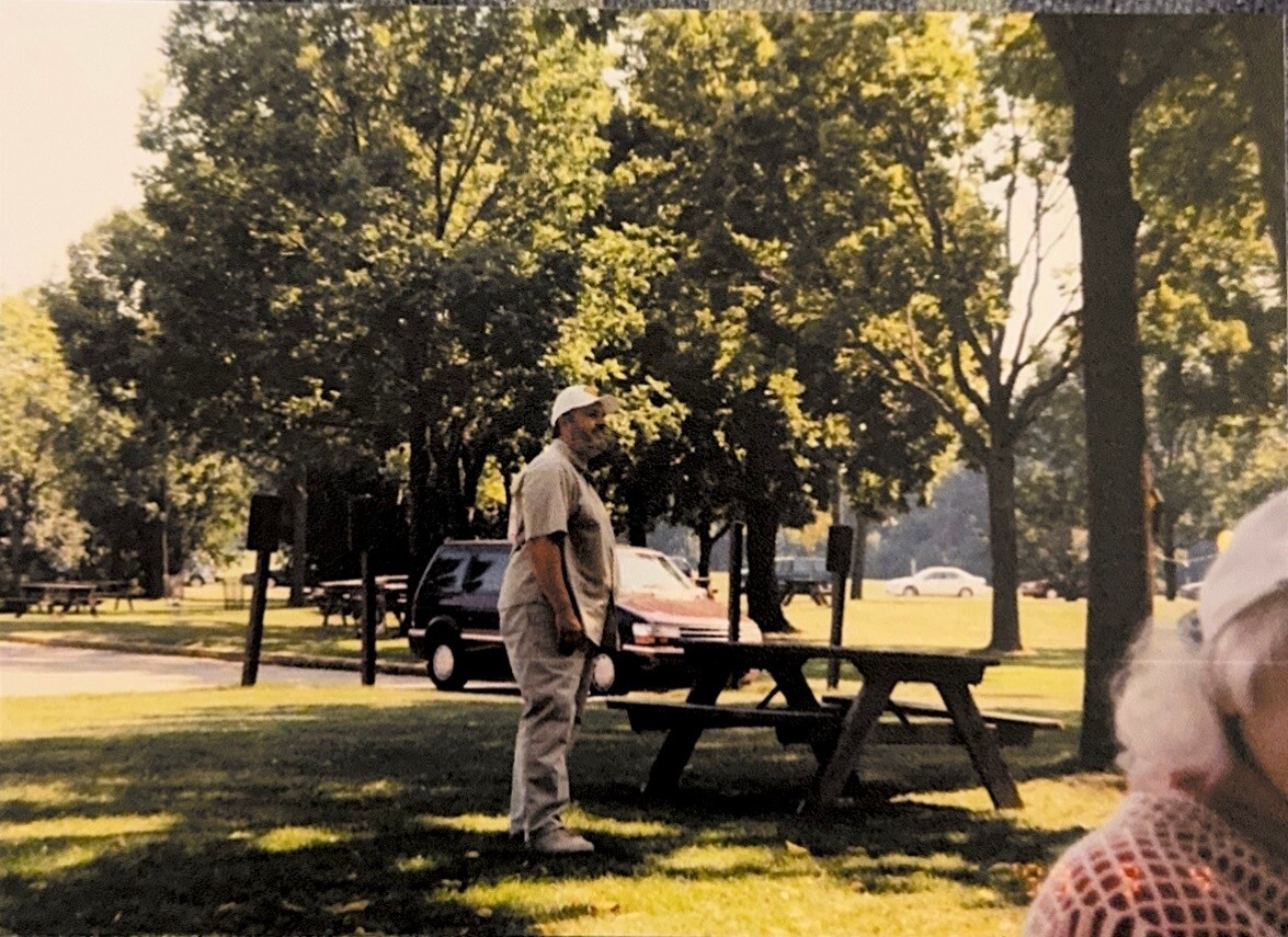 Man standing beside a wooden picnic table in a sunny park, with trees and cars in the background.