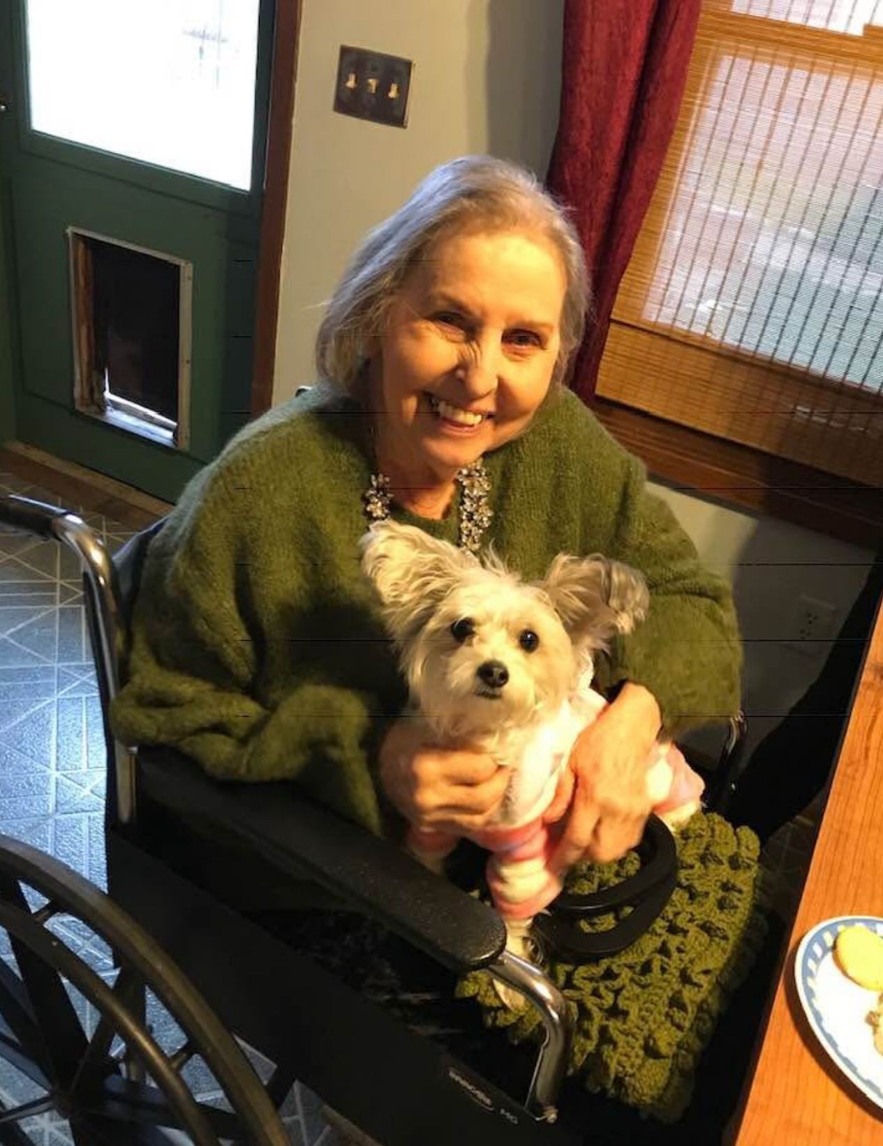 Elderly woman in a green sweater smiling while sitting in a wheelchair, holding a small white fluffy dog in a pink sweater.