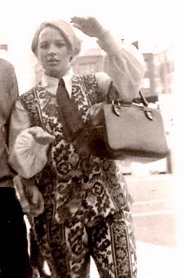 Person in a vintage patterned suit with a tie, adjusting hair as they walk on a city street, carrying a shoulder bag.
