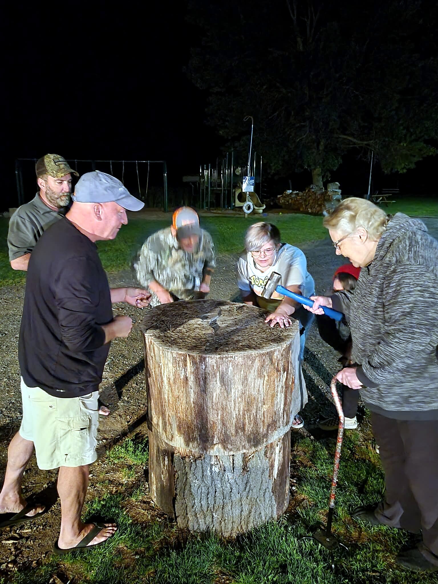 People gathered around a large tree stump at night, chiseling or hammering wood with a blue-handled tool.