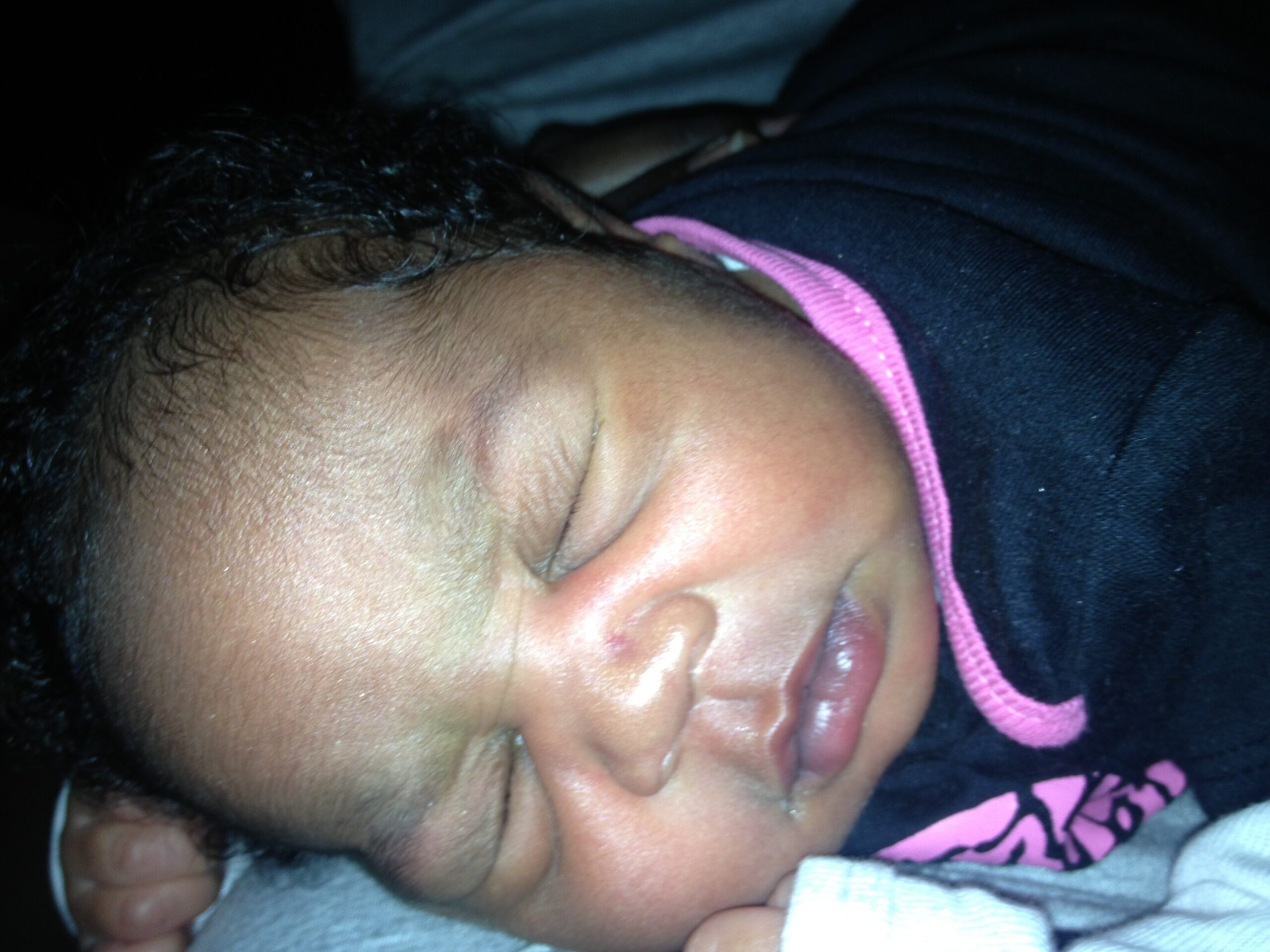Close-up of a sleeping baby with dark hair wearing a pink garment on a dark background.
