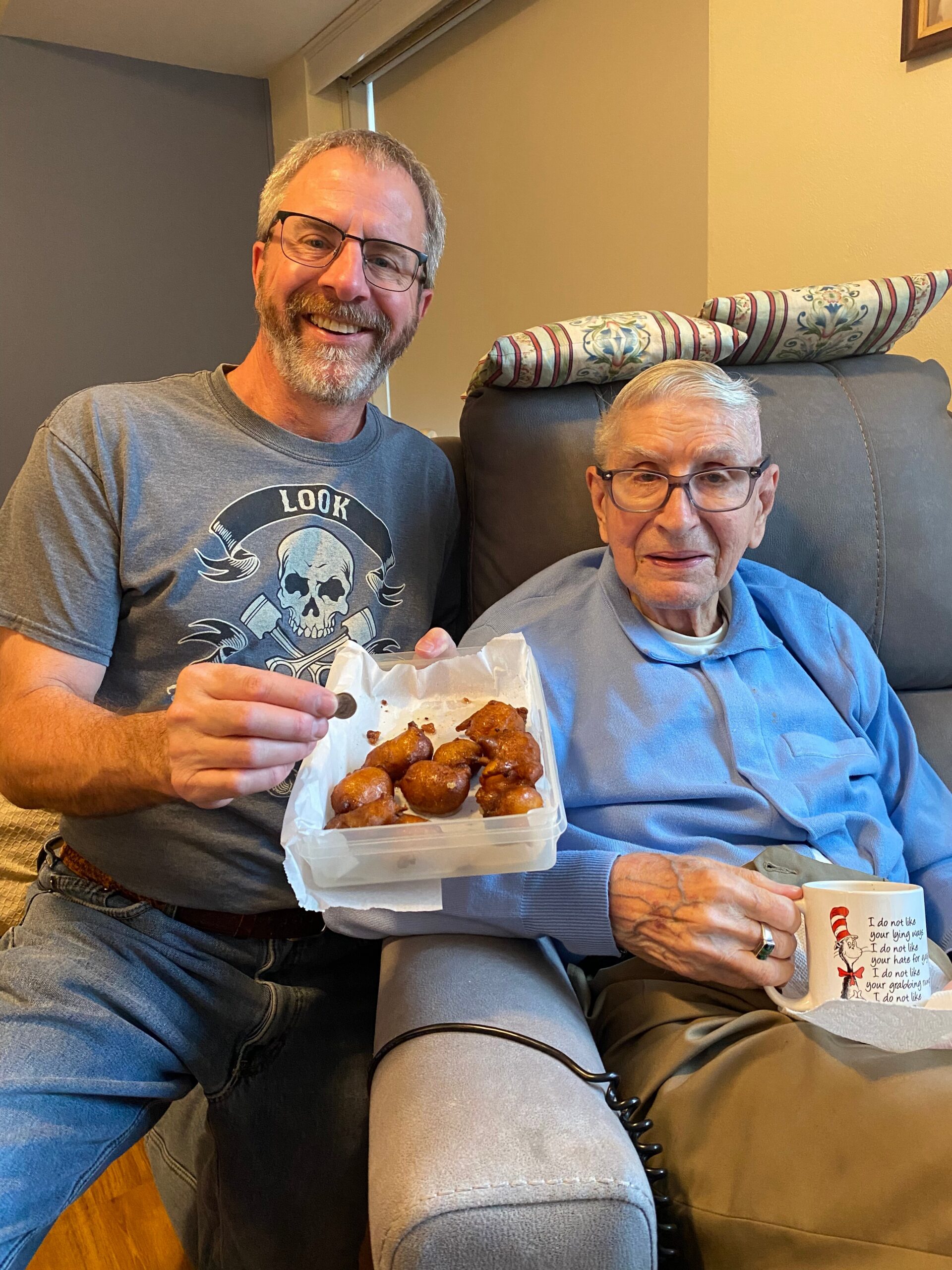 Two smiling men sit on a couch; the man on the left holds a takeout container of fried chicken pieces.