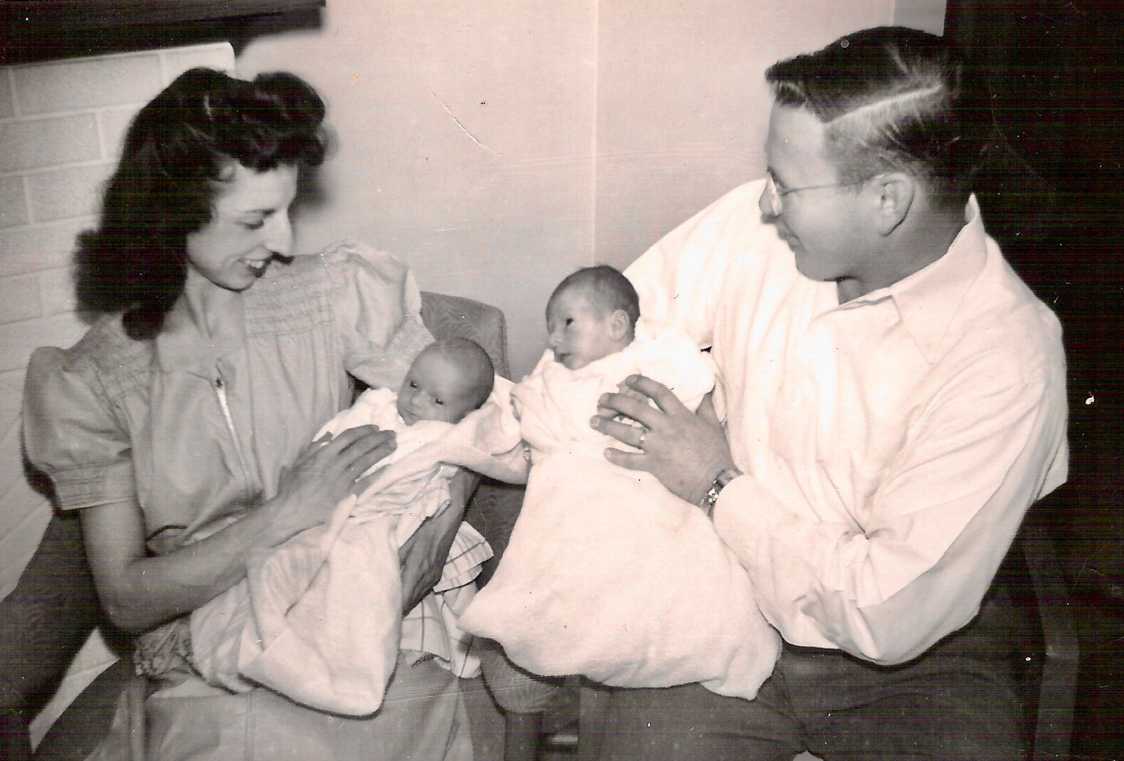 Mother and father cradle twin newborns swaddled in blankets, smiling at their babies in a cozy setting.
