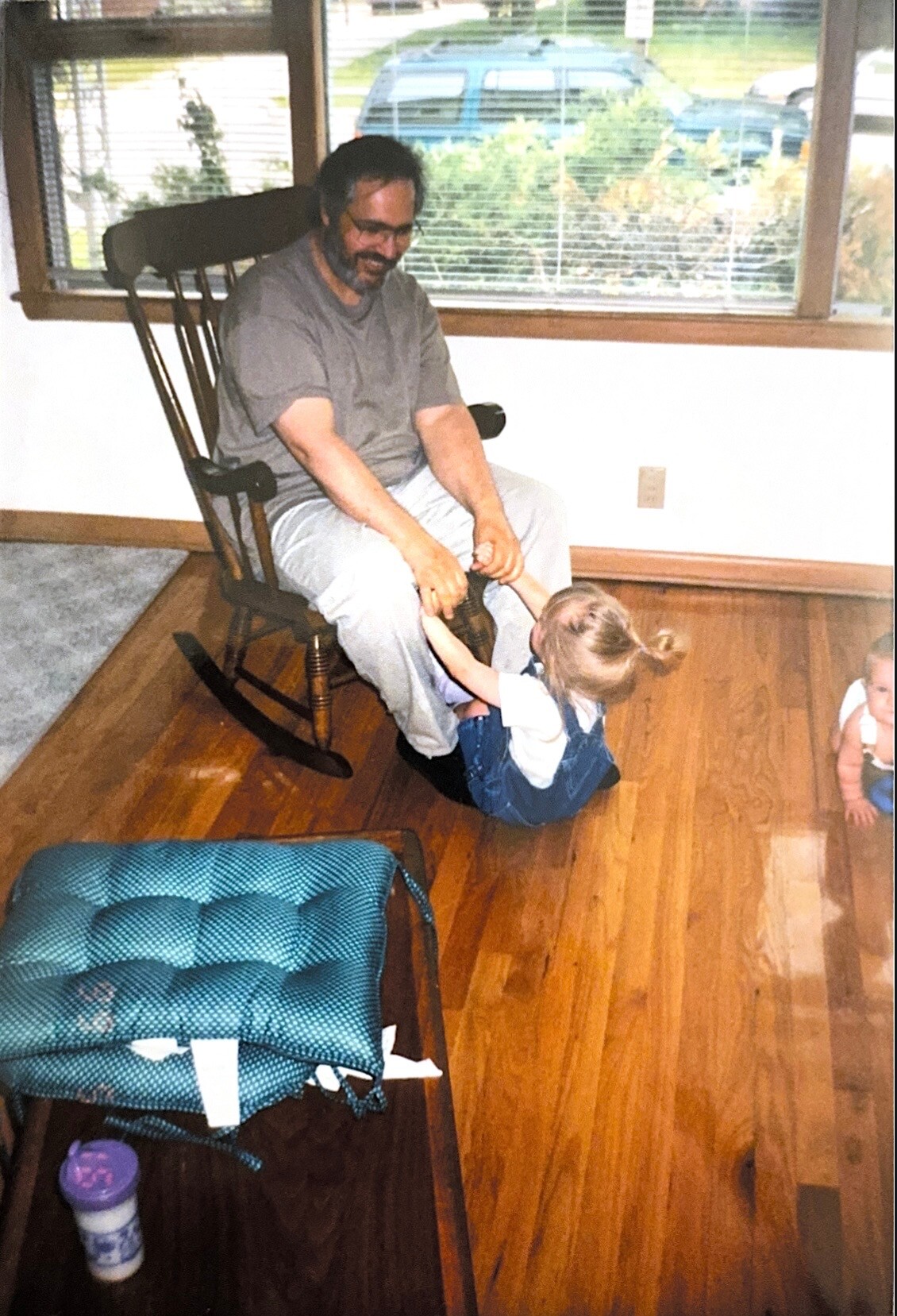 Man in a gray t-shirt sits in a wooden rocking chair, smiling, as a young child sits on the floor and pulls his hands. Two other kids are nearby in a sunny living room.