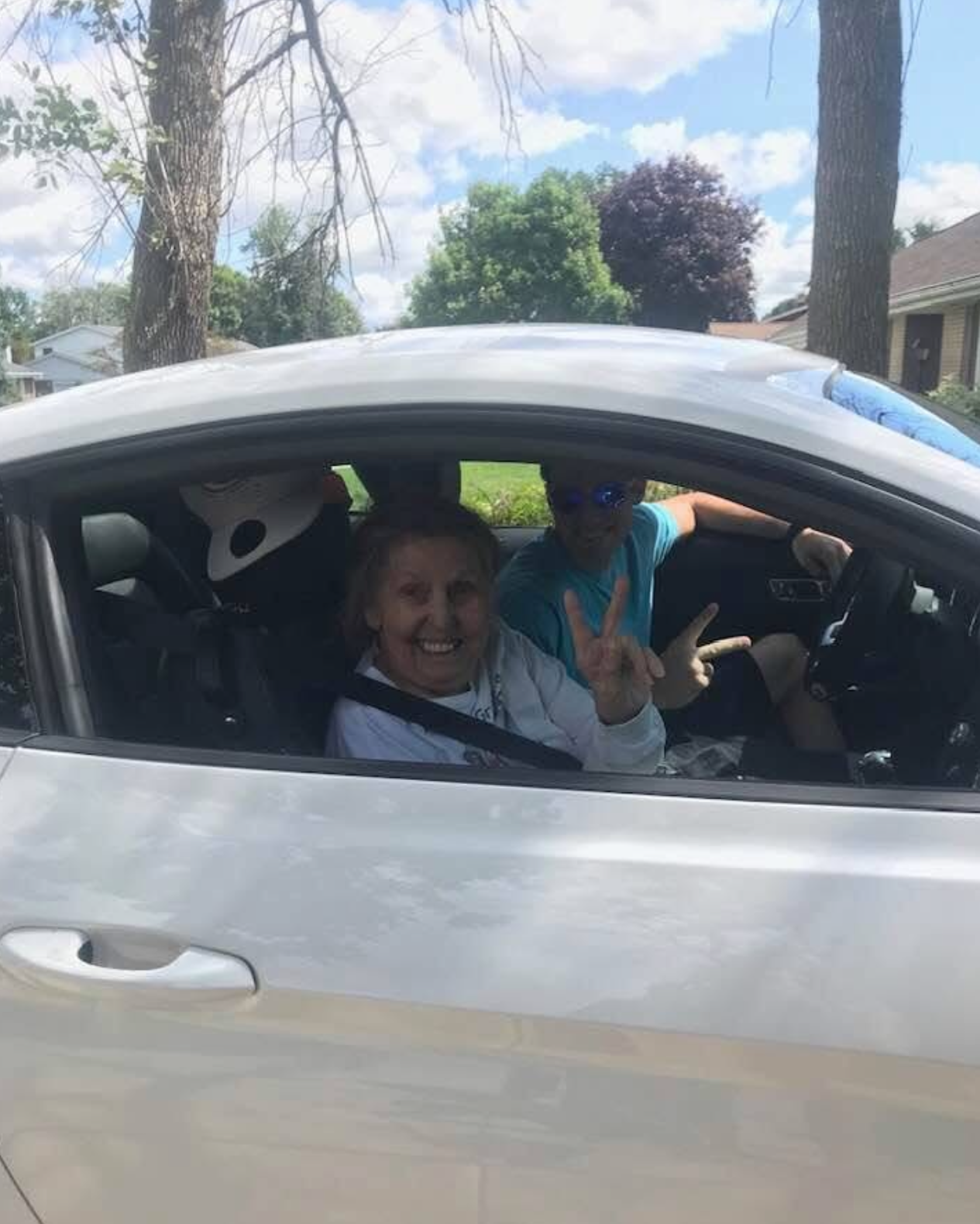Smiling woman in the front seat wearing a seatbelt, two people in the back making peace signs through the window in a silver car on a sunny day