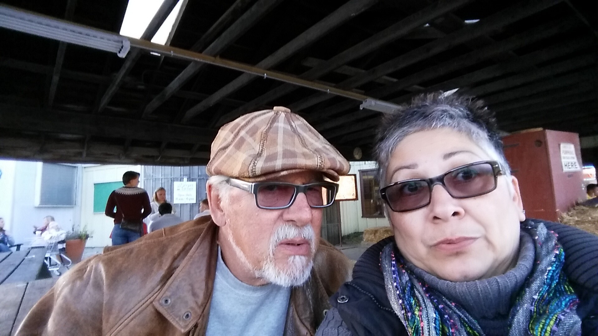 Two people take a close-up selfie under a dark wooden pavilion, with other diners in the background.