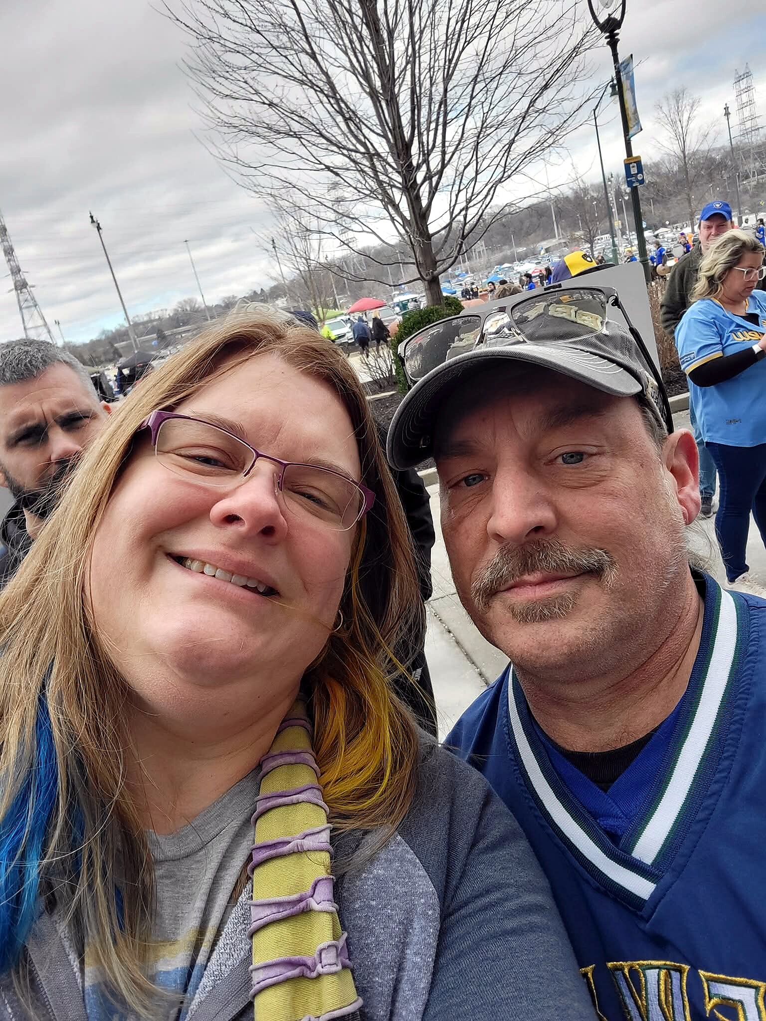 Close-up selfie of a smiling woman with glasses and a man in a blue jacket at an outdoor event; crowd and tents in the background on a cloudy day.