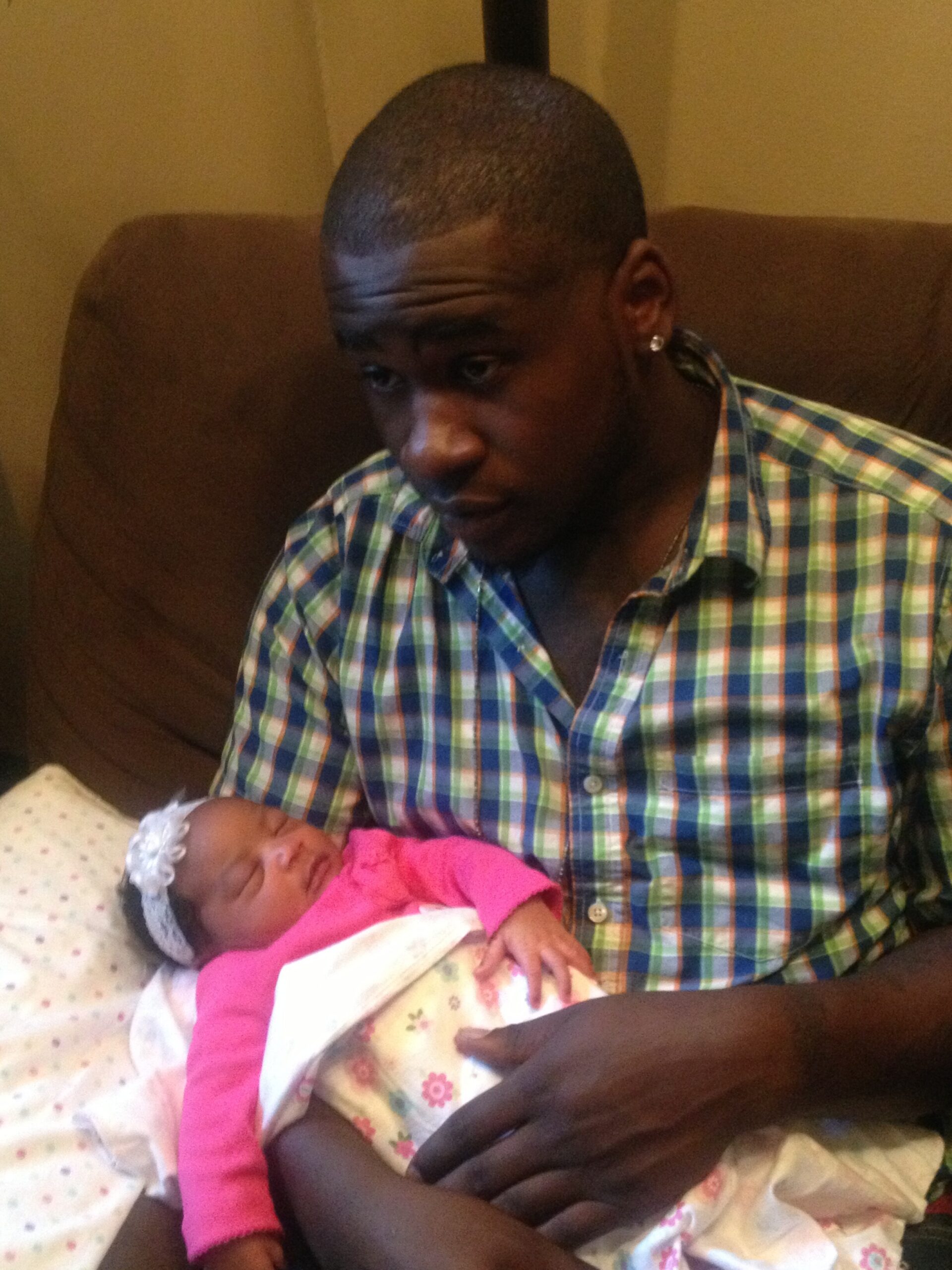 Father with a short haircut sits on a brown couch, cradling a newborn baby wrapped in a floral blanket and pink clothing.