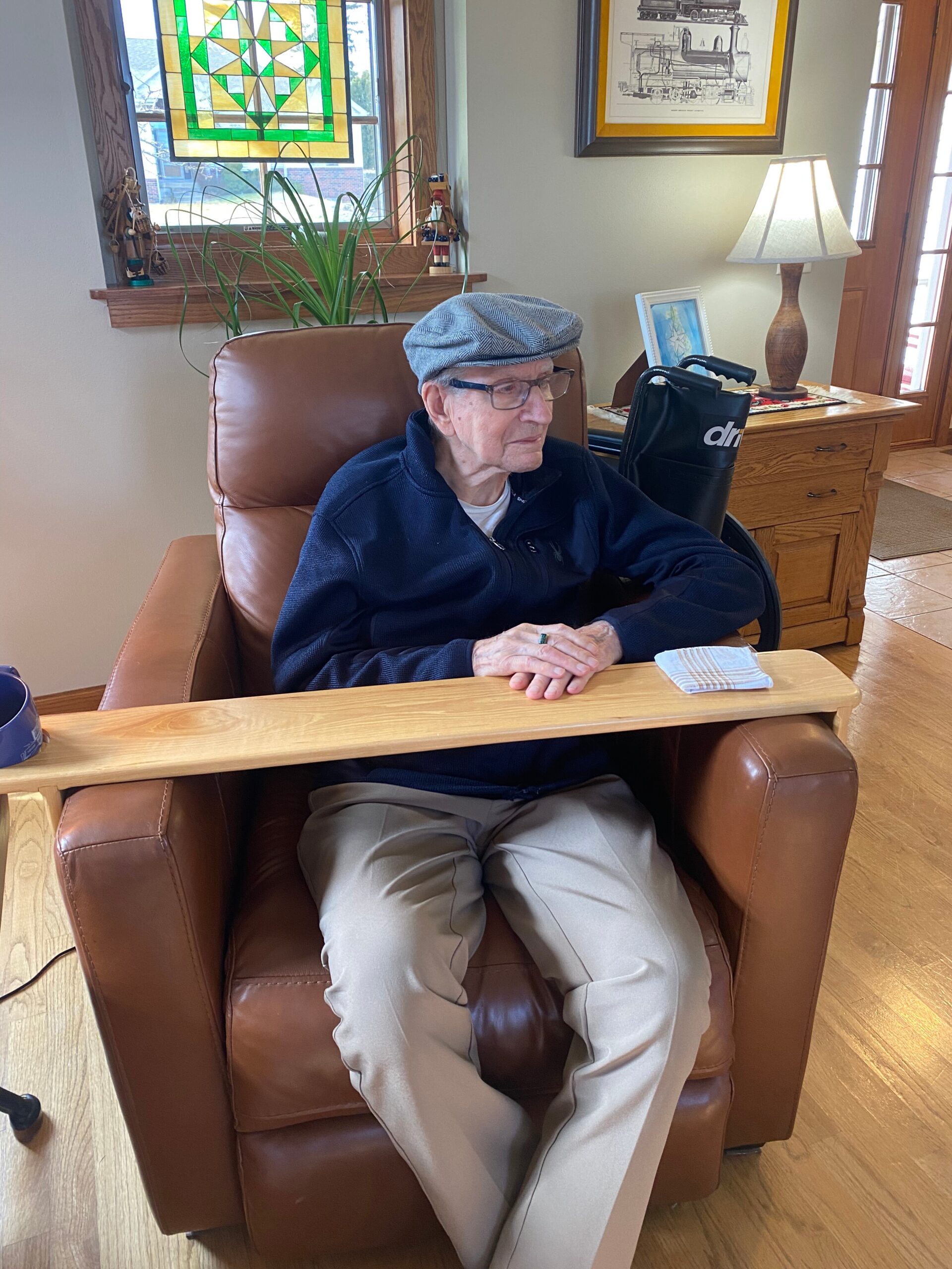 Senior man wearing a flat cap and glasses sits in a brown leather chair at a wooden table in a cozy living room with a stained-glass window behind him.
